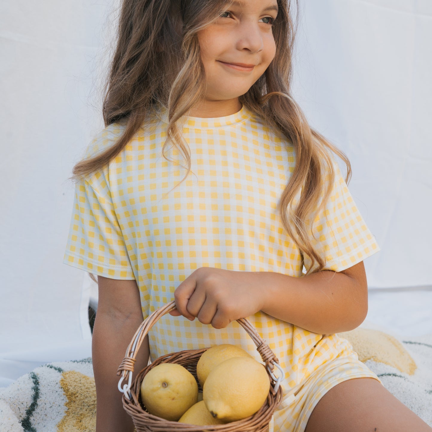 A smiling young girl with long brown hair wears the forever french baby Girl's Bamboo Short Set | Citron and holds a small basket of lemons, sitting against a white fabric background.