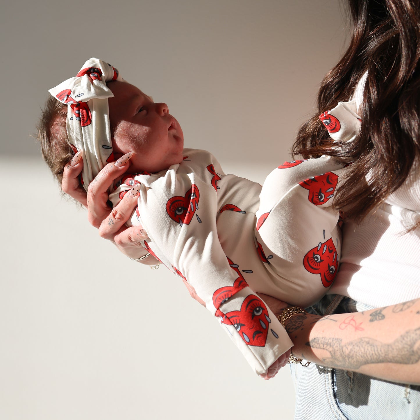 A woman holds a baby dressed in forever french baby's Bamboo Zip Pajamas | Sad Girl—a white outfit with red heart designs. Her tattooed arm catches the sunlight as they pose against a plain background.