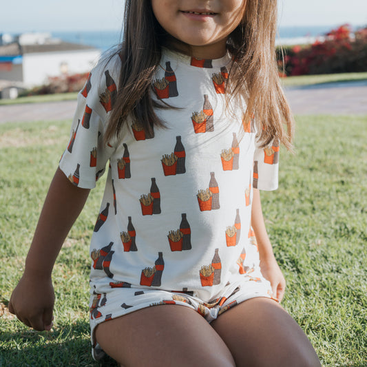 A young girl sits on grass, wearing the forever french baby Girl’s Bamboo Short Set | Fries & Cola—white T-shirt and shorts printed with soda bottles and red French fries cartons. Greenery and distant buildings blur in the background.