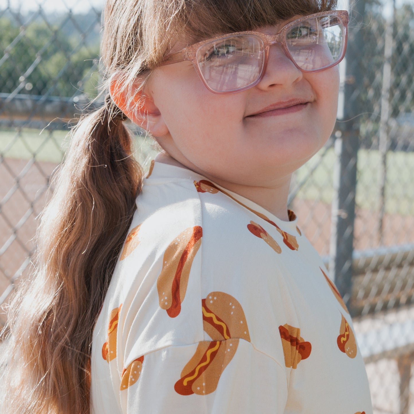 A young girl with long brown hair in a ponytail, wearing large glasses and the Girl's Bamboo Short Set | Hot Dogs by forever french baby, smiles softly by a chain-link fence at a baseball field.
