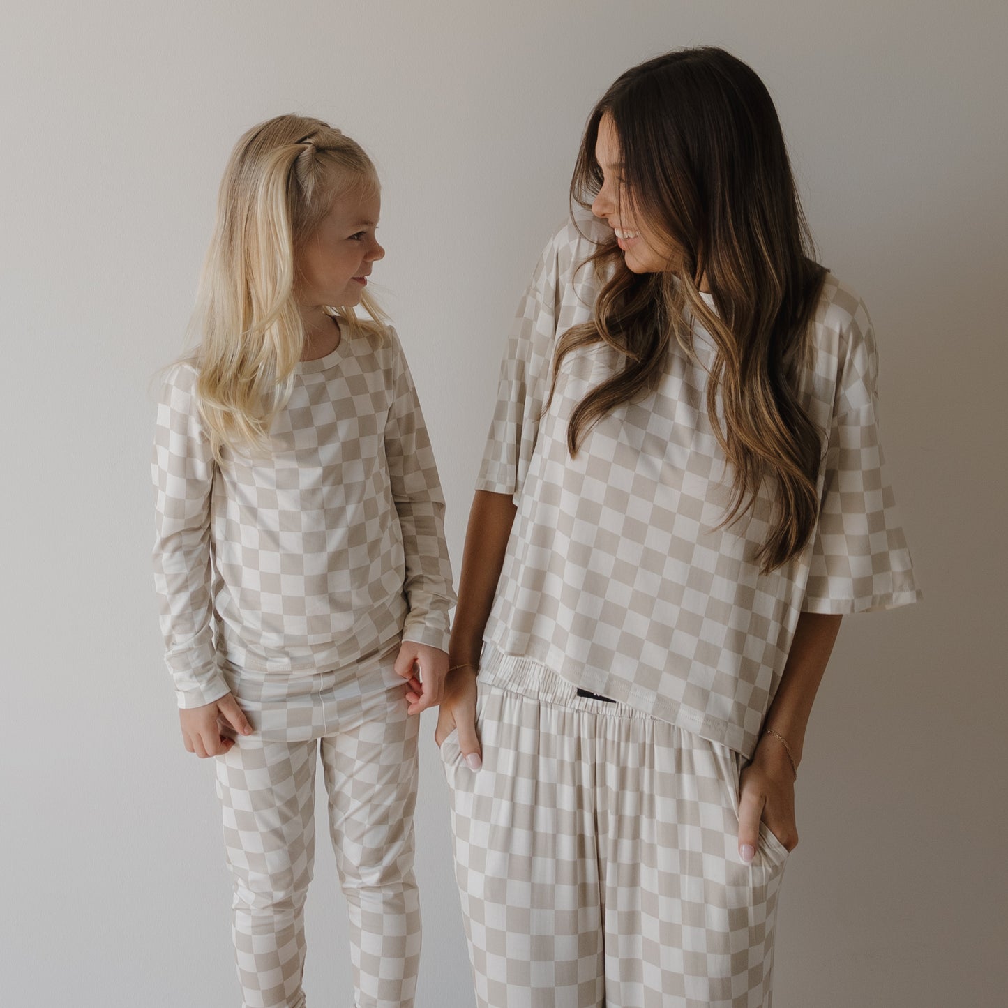 A woman and a young girl smile at each other, both wearing matching forever french baby Women's Boxy Tee Lounge Sets in Checkerboard, in a minimal, well-lit setting with a plain light background.