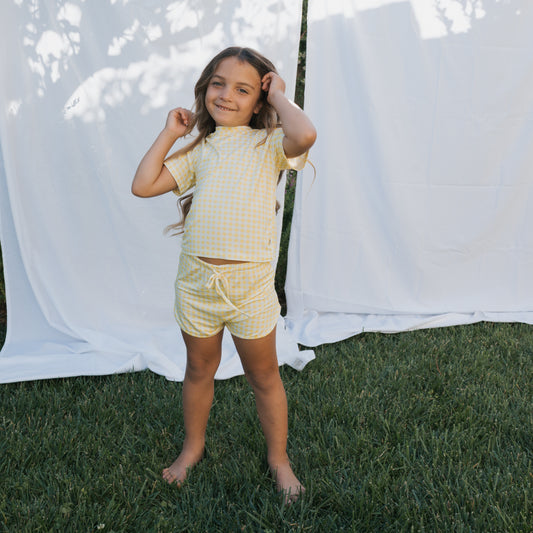 A young girl stands barefoot on grass, smiling and holding her long hair. She wears the forever french baby Girl's Bamboo Short Set in Citron, with a white fabric backdrop outdoors.