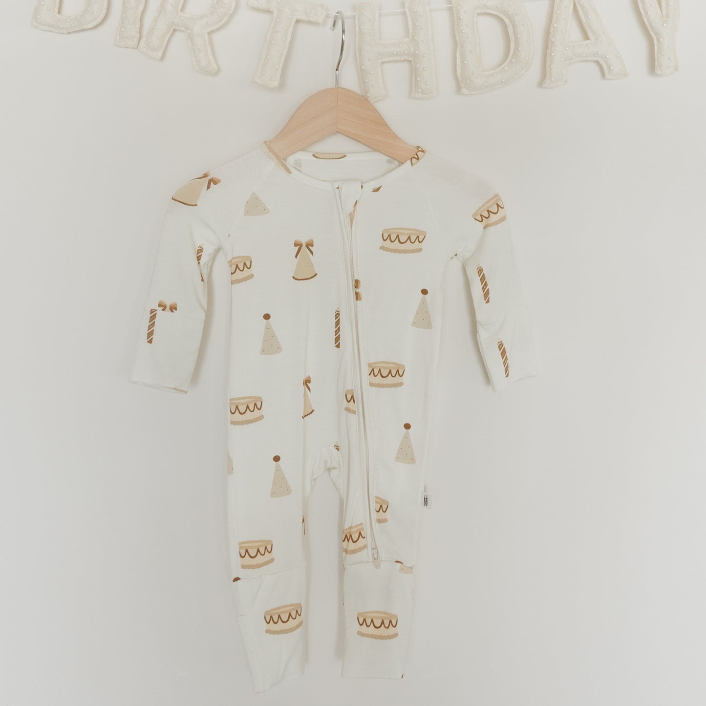 A white Bamboo Zip Pajama from forever french baby’s “Birthday Girl” collection, adorned with cheerful cake, candles, and party hat prints, hangs on a wooden hanger. Crafted from hypoallergenic materials gentle on sensitive skin, this footie pajama offers all-day comfort. Above it, a fabric “BIRTHDAY” banner is partially visible.