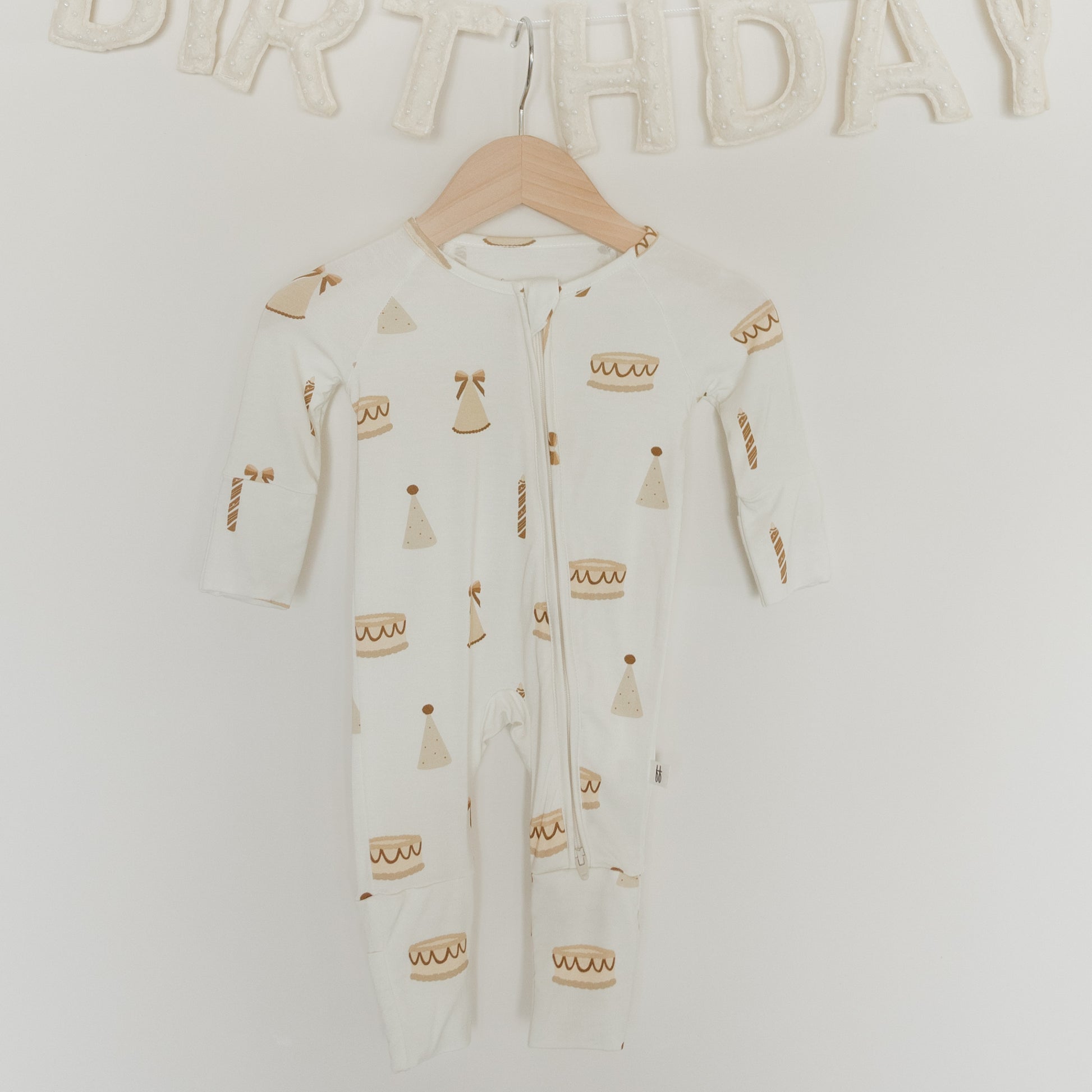 A white Bamboo Zip Pajama from forever french baby’s “Birthday Girl” collection, adorned with cheerful cake, candles, and party hat prints, hangs on a wooden hanger. Crafted from hypoallergenic materials gentle on sensitive skin, this footie pajama offers all-day comfort. Above it, a fabric “BIRTHDAY” banner is partially visible.