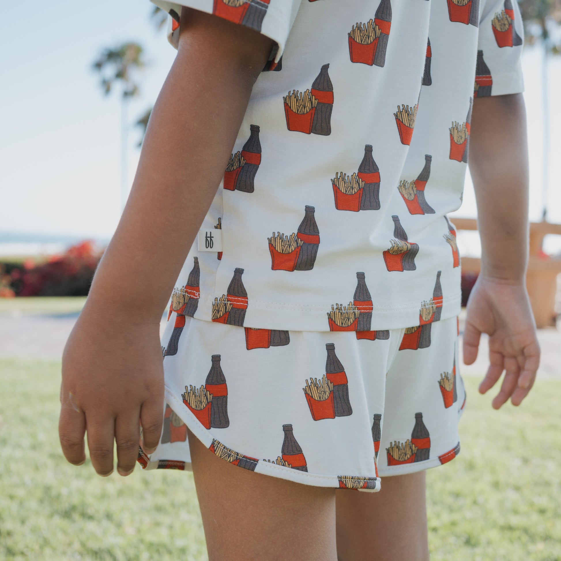 A child wears the forever french baby Girl's Bamboo Short Set in Fries & Cola print, featuring soda bottles and fries, while standing outdoors on grass with palm trees and a blue sky in the background.