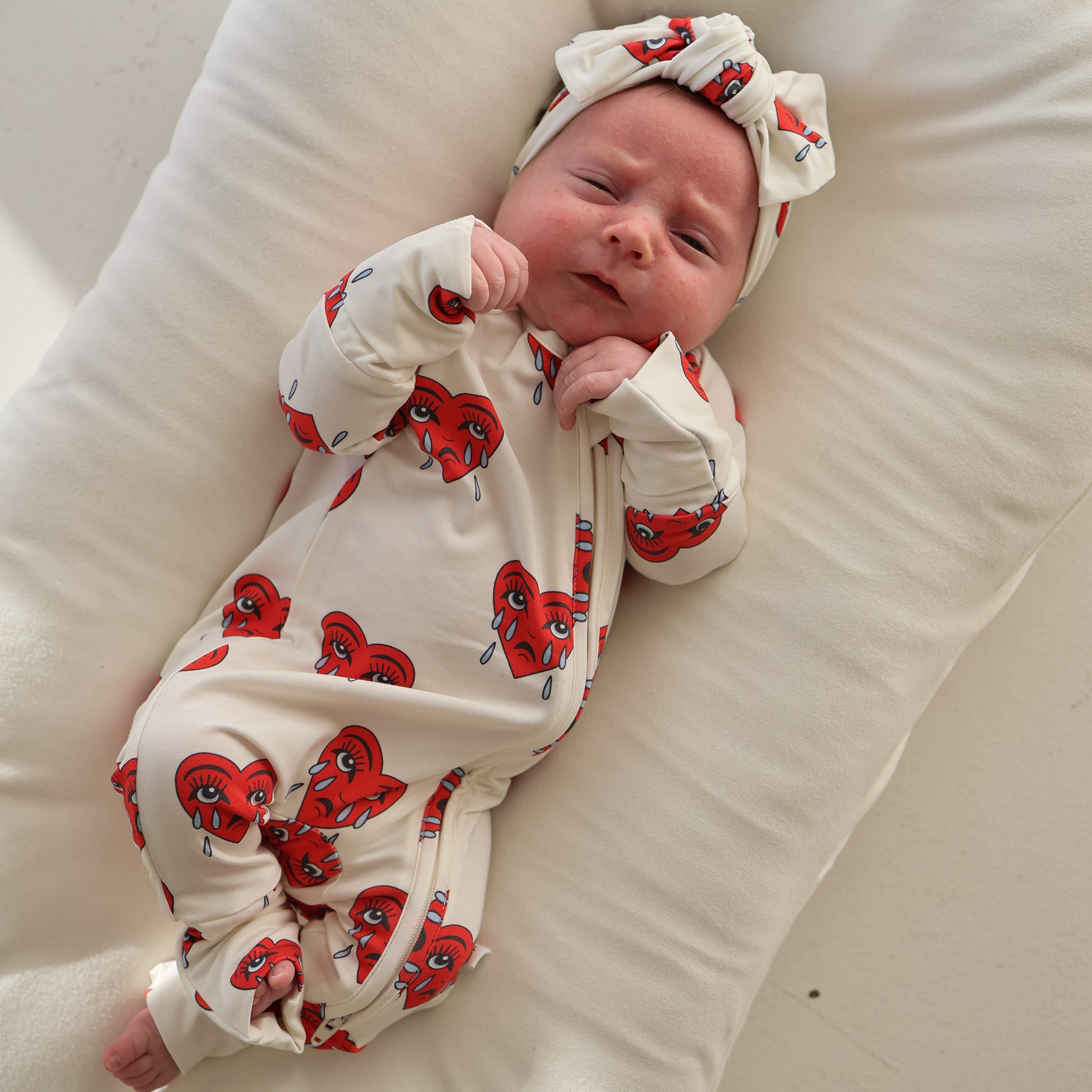 A newborn in forever french baby’s Bamboo Zip Pajamas | Sad Girl—white with red heart and eye prints—lies on a soft, white cushion, hands near face and eyes partly closed.