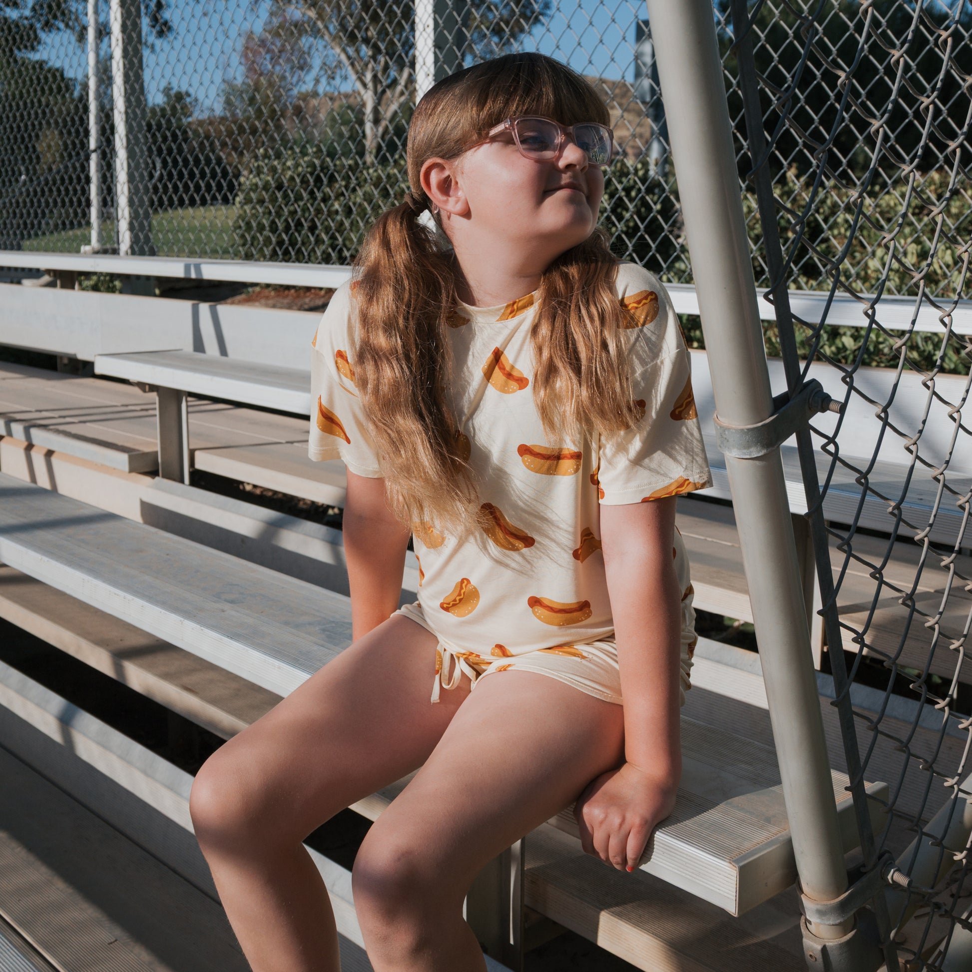 A girl with long light brown pigtails and glasses sits on bleachers, wearing the forever french baby Girl's Bamboo Short Set | Hot Dogs, as sunlight filters through a chain-link fence behind her.