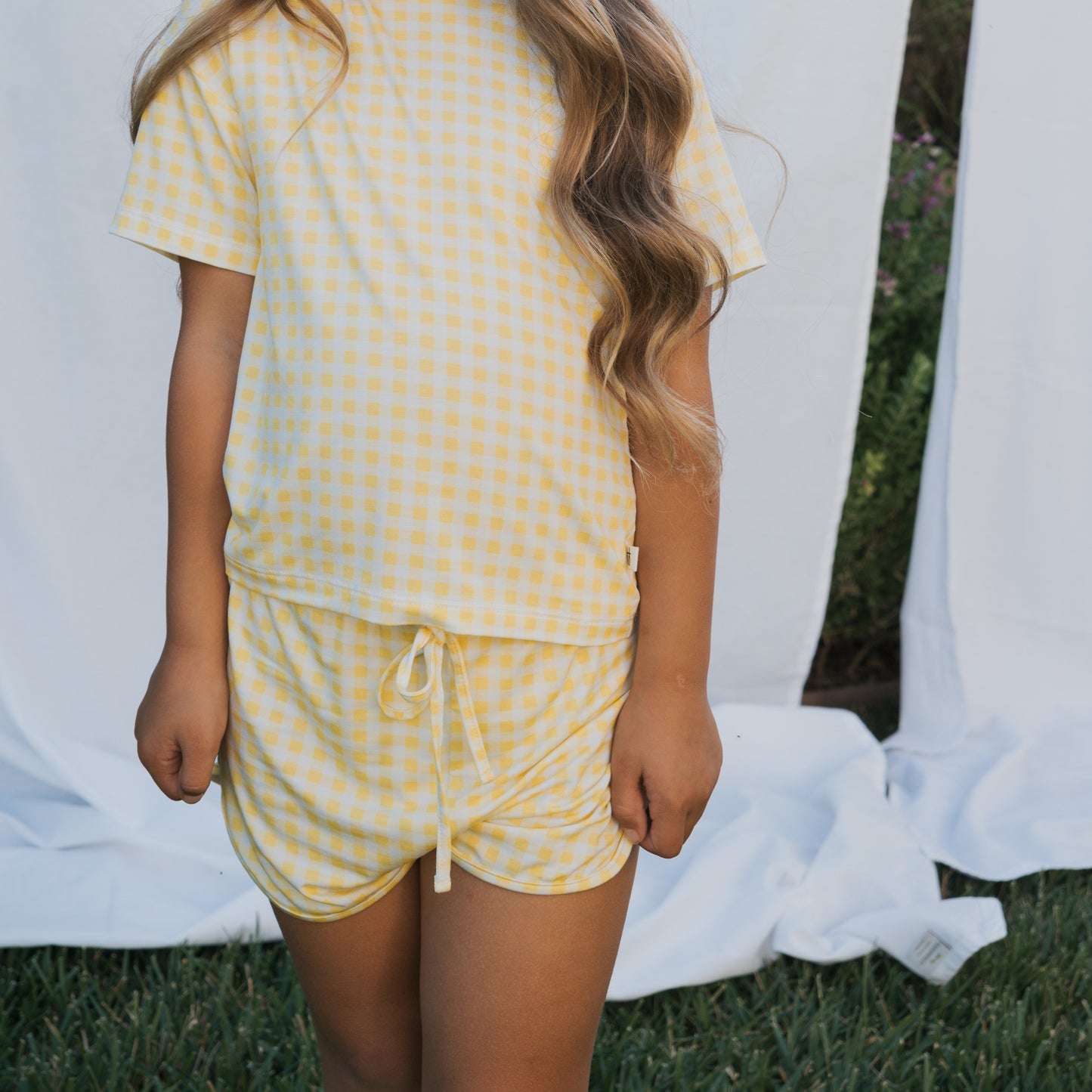 A child wears the forever french baby Girl's Bamboo Short Set | Citron, a breathable yellow and white checkered short-sleeve shirt with matching shorts. Their long wavy hair falls over one shoulder, soft white fabric in the background.