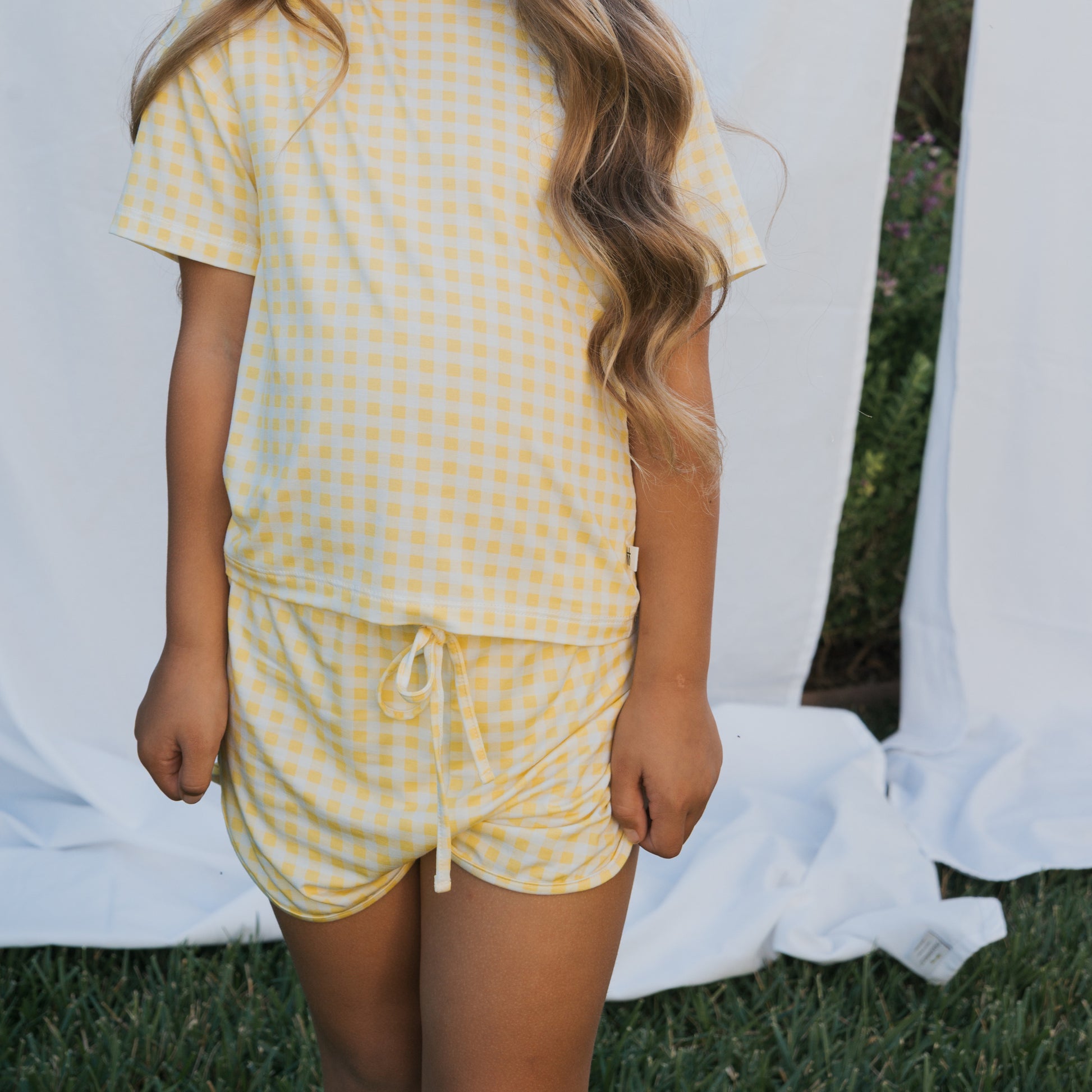 A child wears the forever french baby Girl's Bamboo Short Set | Citron, a breathable yellow and white checkered short-sleeve shirt with matching shorts. Their long wavy hair falls over one shoulder, soft white fabric in the background.