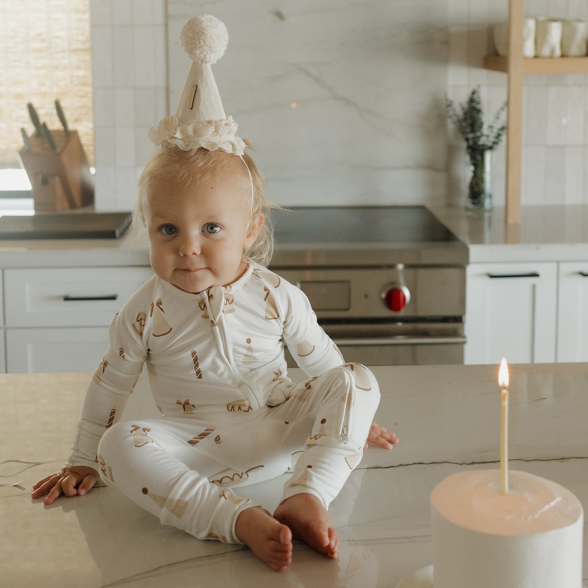 A toddler wearing a party hat and forever french baby Bamboo Zip Pajamas | Birthday Girl sits on a kitchen counter, looking at the camera. In the foreground, a white candle with a lit flame is visible.