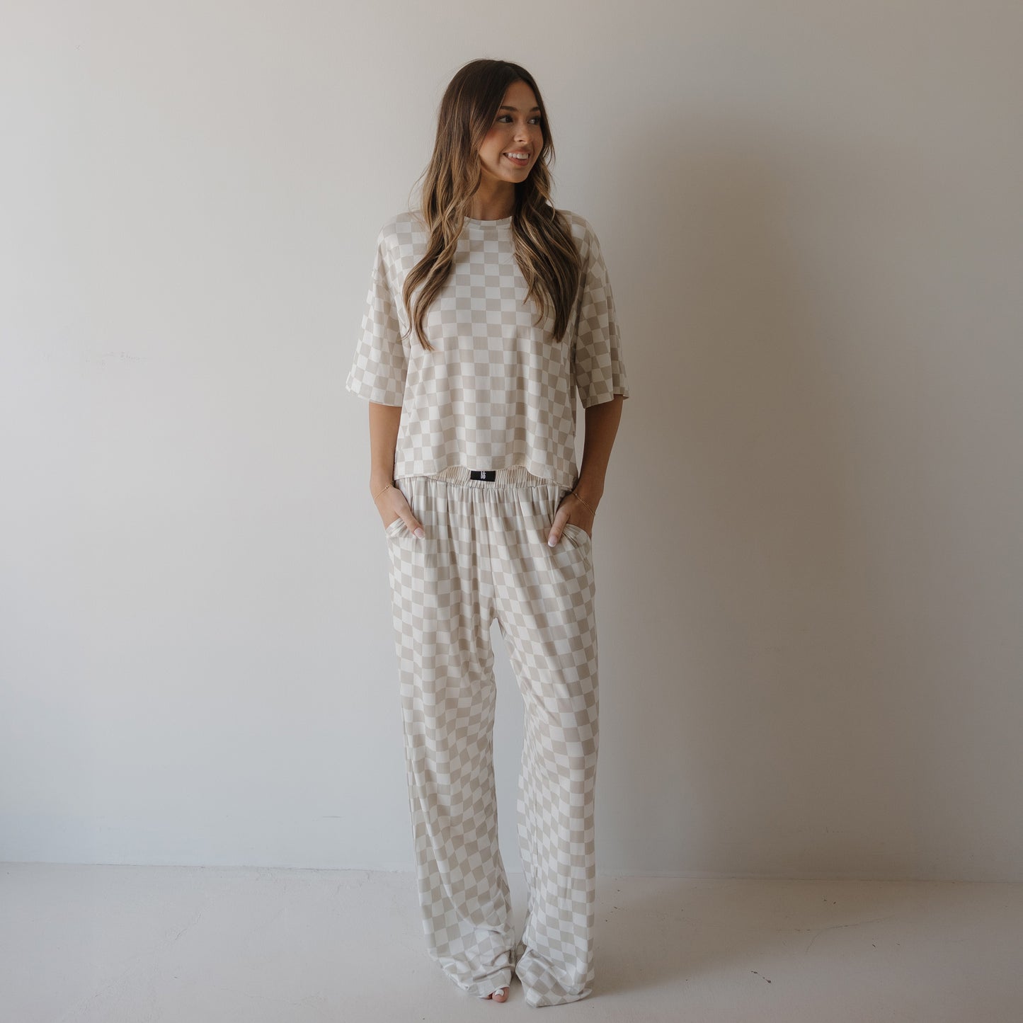 A woman with long, wavy hair stands against a white wall wearing the forever french baby Women's Boxy Tee Lounge Set | Checkerboard, featuring a beige and white checkered pattern, short sleeves, and loose-fitting pants with her hands in her pockets.