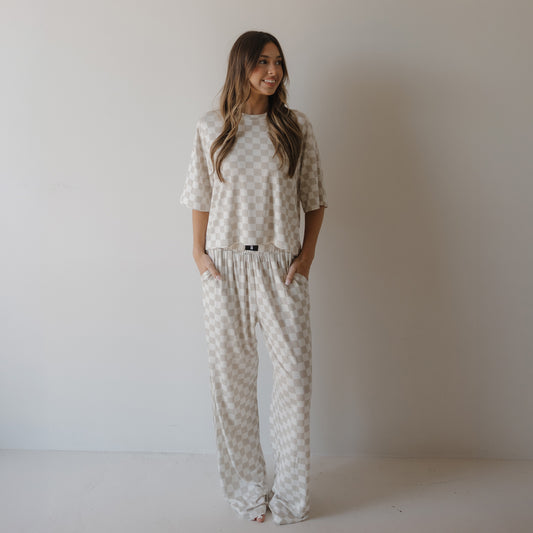 A woman with long, wavy hair stands against a white wall wearing the forever french baby Women's Boxy Tee Lounge Set | Checkerboard, featuring a beige and white checkered pattern, short sleeves, and loose-fitting pants with her hands in her pockets.