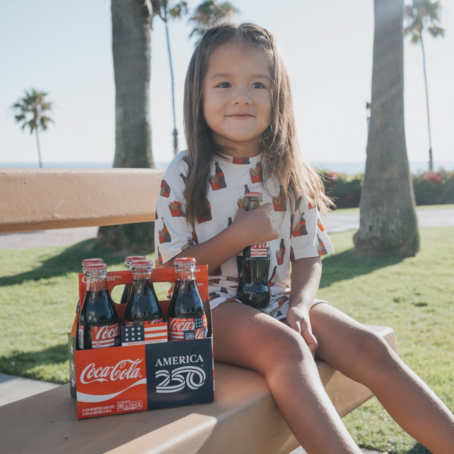 A young girl in the forever french baby Girl's Bamboo Short Set | Fries & Cola smiles on a park bench, holding a Coca-Cola bottle beside an "America 250" six-pack. Palm trees and grass fill the background.