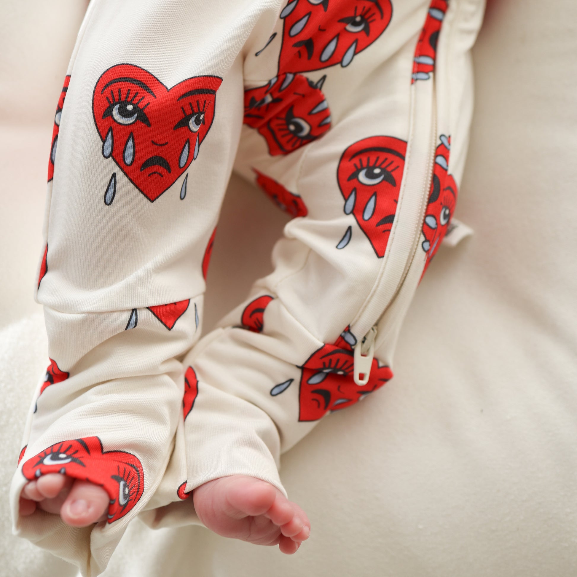 A baby wearing forever french baby's Bamboo Zip Pajamas | Sad Girl, featuring a cream color with red crying heart designs and rollover cuffs, lies on a soft white surface with only their legs and part of an arm visible.