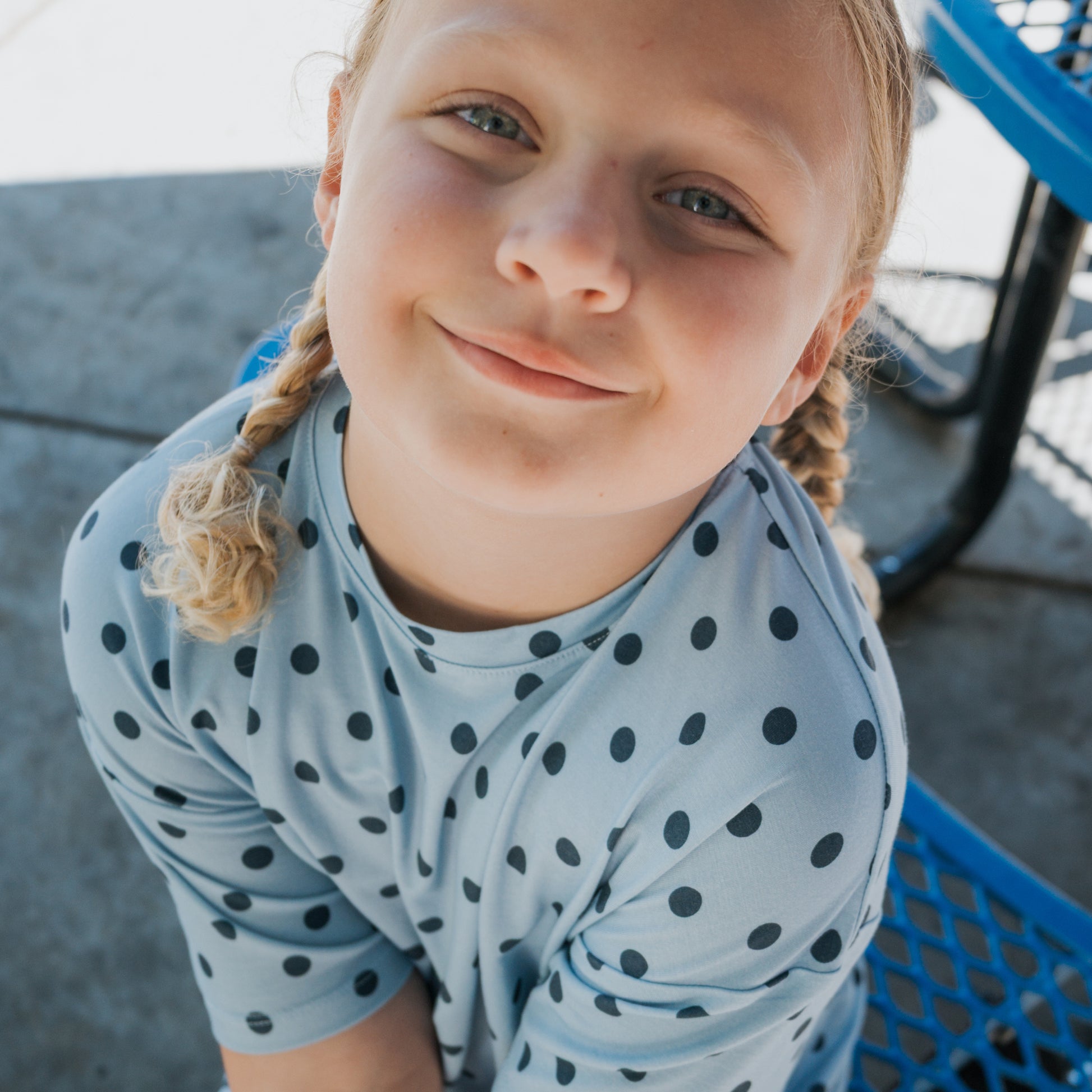 A smiling child with blonde braided hair sits outdoors on a blue metal bench, wearing the forever french baby Girl's Bamboo Short Set | Sully—an airy, light blue outfit with black polka dots.