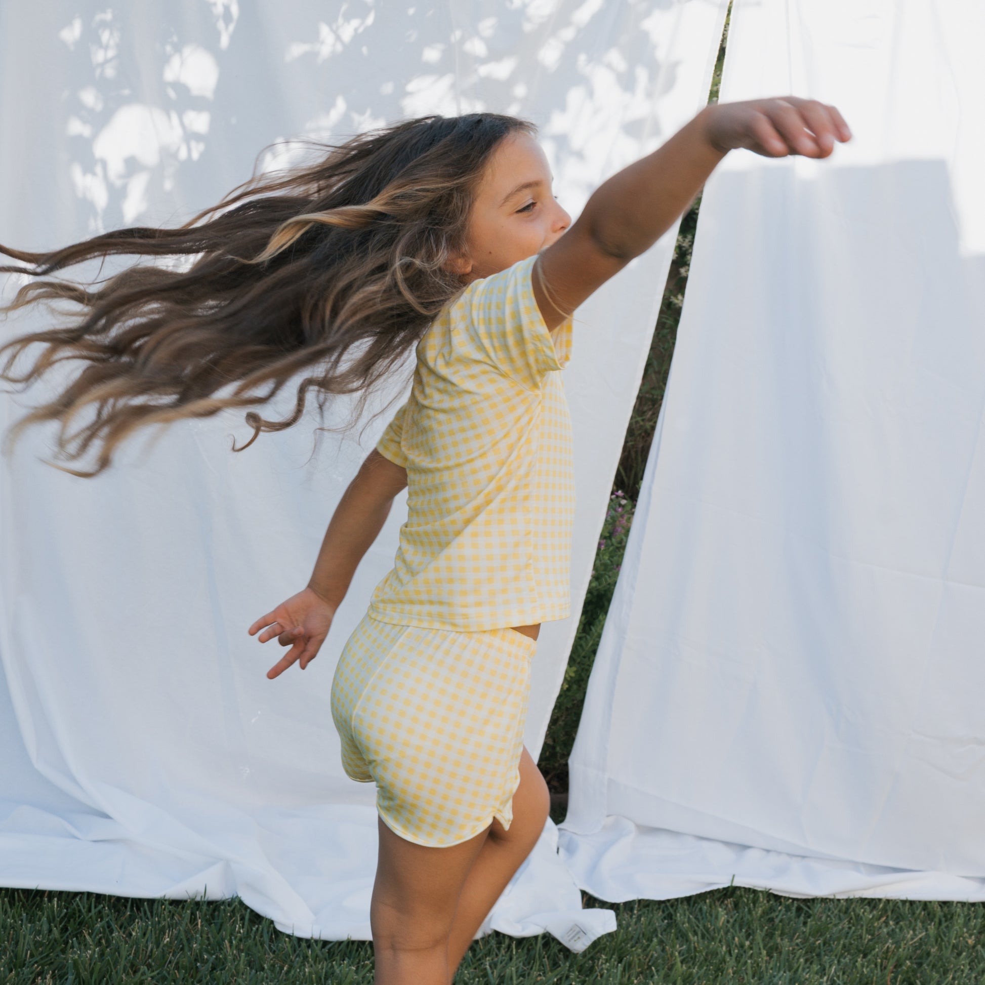 A young girl with long brown hair twirls on grass, wearing the forever french baby Girl's Bamboo Short Set | Citron—breathable yellow checkered shorts and matching t-shirt—with her arm raised before a white backdrop.