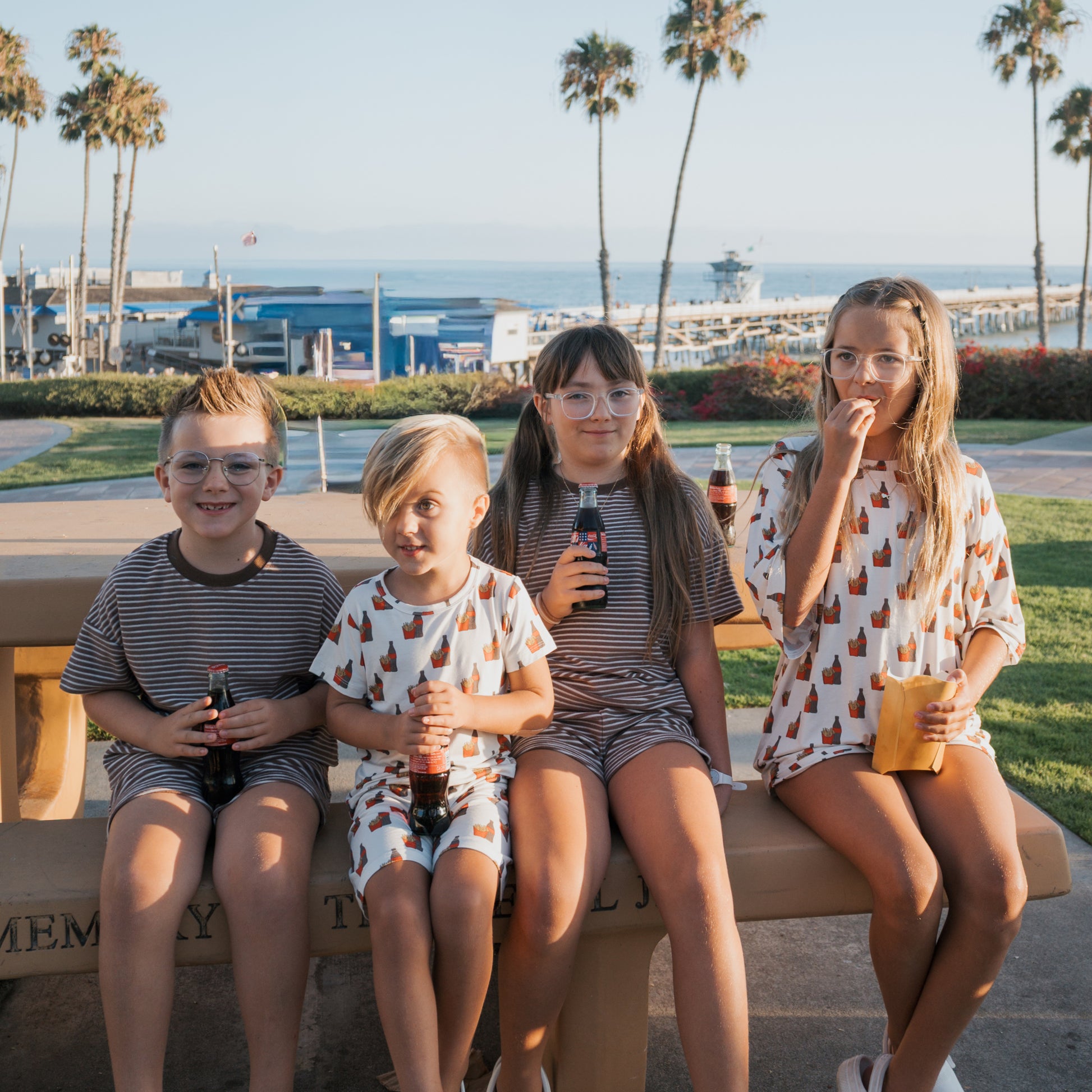 Four girls in forever french baby Girl's Bamboo Short Sets featuring Fries & Cola prints sit on a beachside bench with Coke and juice. Palm trees, grass, and the ocean glimmer in the sunlight behind them.