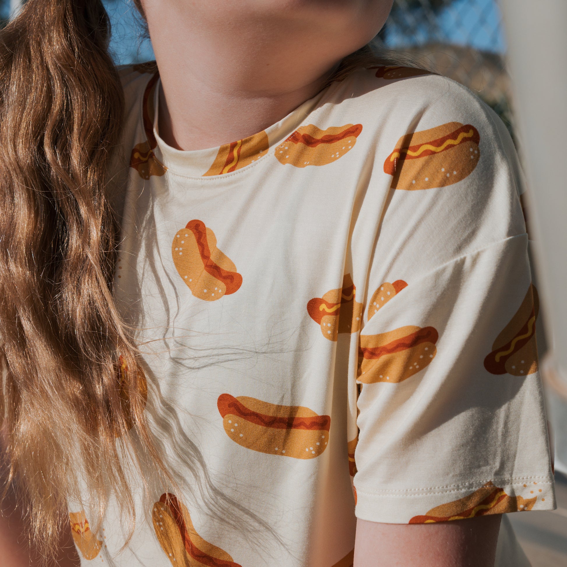 A girl with long, wavy hair wears the forever french baby Girl's Bamboo Short Set in Hot Dogs print, featuring a cream-colored, hypo-allergenic T-shirt while sitting in sunlight.