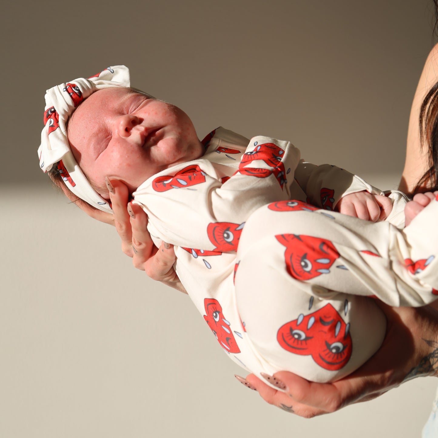 A baby in forever french baby's Bamboo Zip Pajamas | Sad Girl—a white hypoallergenic outfit with red cartoon faces—is gently held in someone's arms against a plain, sunlit background.