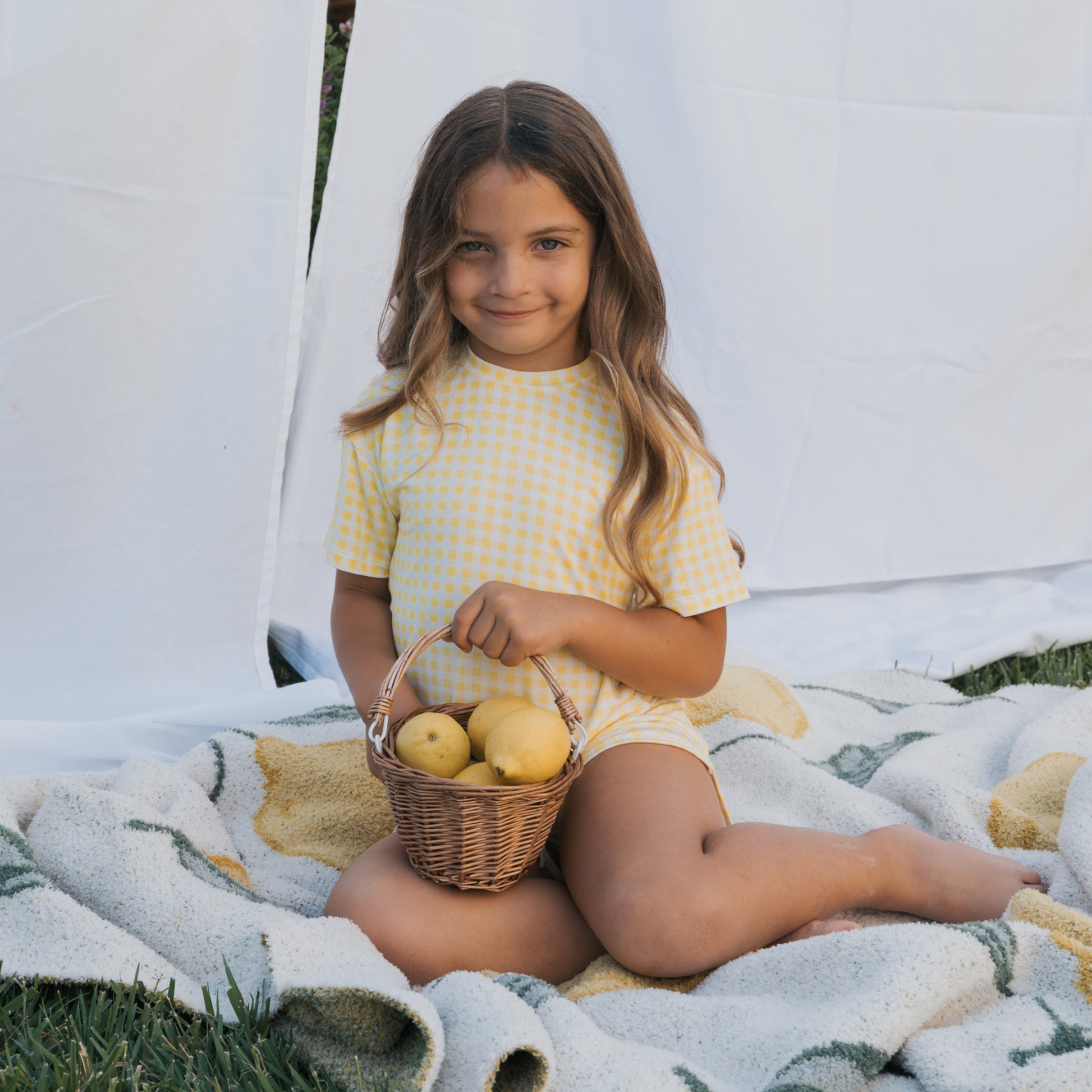 A young girl with long brown hair smiles outdoors, sitting on a blanket with a basket of lemons. She wears the forever french baby Girl's Bamboo Short Set | Citron, featuring a yellow and white checkered breathable design. White fabric drapes behind her.