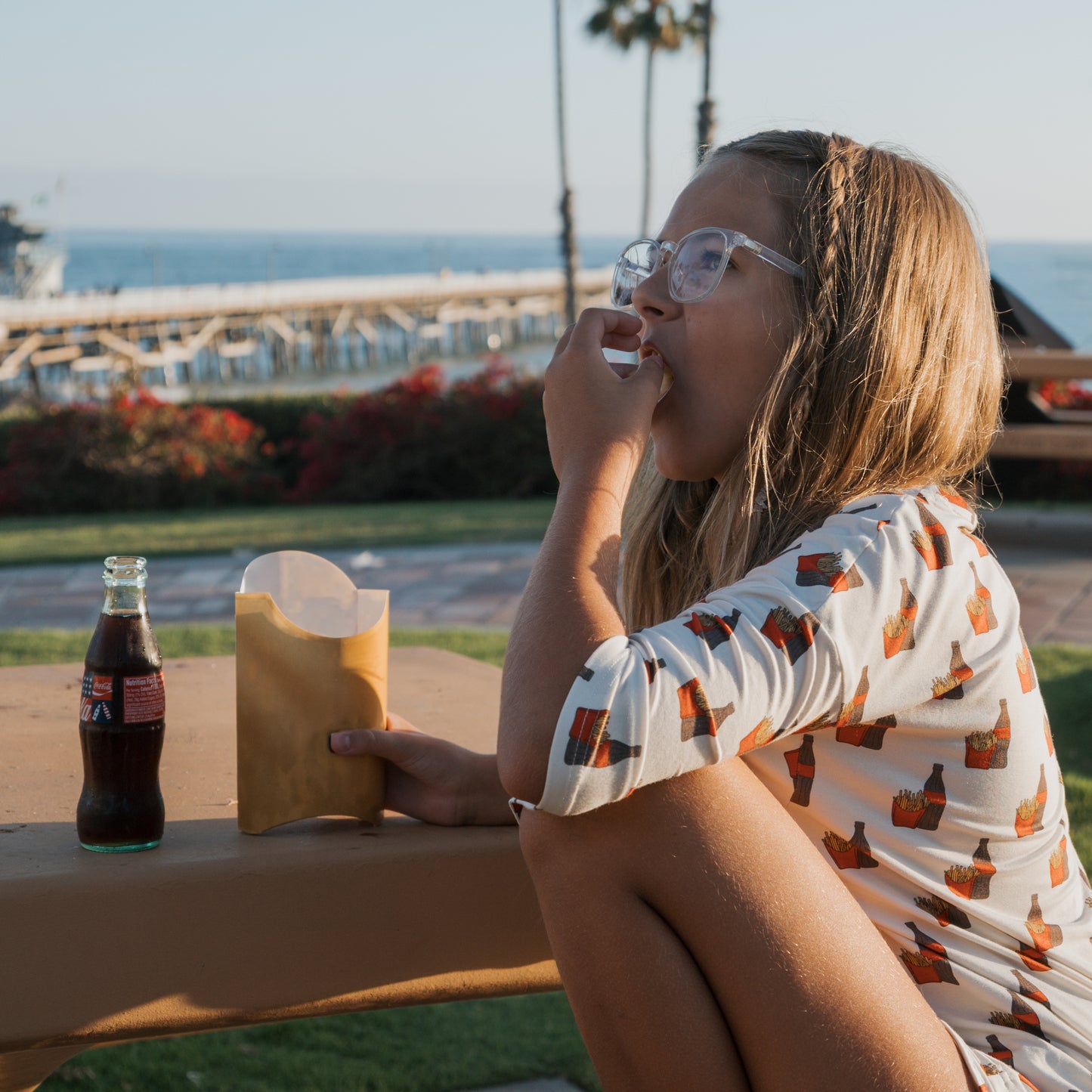 Wearing the forever french baby Girl's Bamboo Short Set | Fries & Cola, a girl with glasses relaxes outdoors by the ocean, enjoying fries and cola amidst a scenic backdrop of a pier and palm trees.