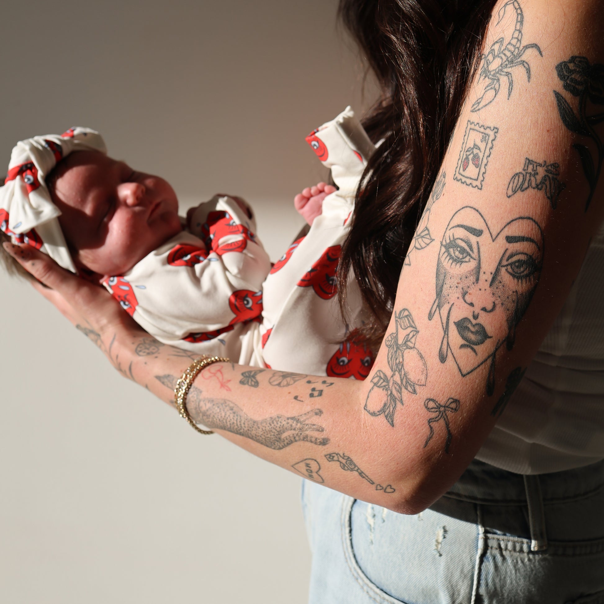 A tattooed woman holds a newborn dressed in forever french baby Bamboo Zip Pajamas | Sad Girl, featuring red patterns and a matching headband. The woman's face is hidden; bright light shines from the left.