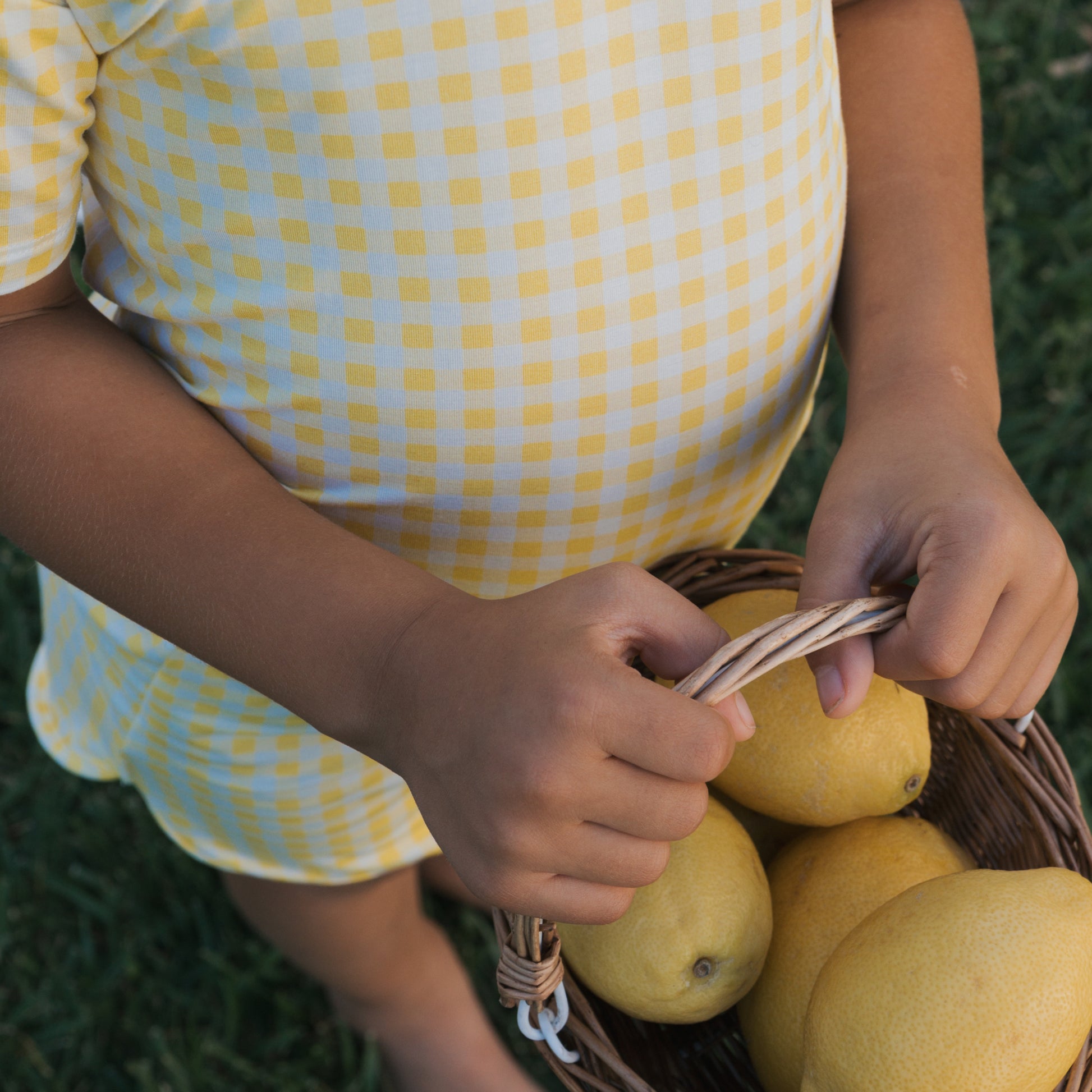 A child wears the forever french baby Girl's Bamboo Short Set | Citron and holds a wicker basket of lemons while standing on green grass.