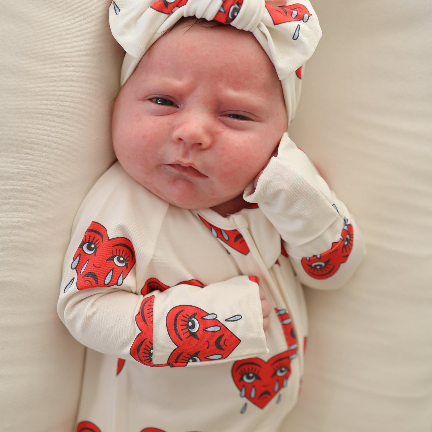 A baby in forever french baby's Bamboo Zip Pajamas | Sad Girl—cream hypoallergenic pajamas with rollover cuffs and a headband featuring red crying heart faces—lies on a cream surface, gazing at the camera with one hand near their face.