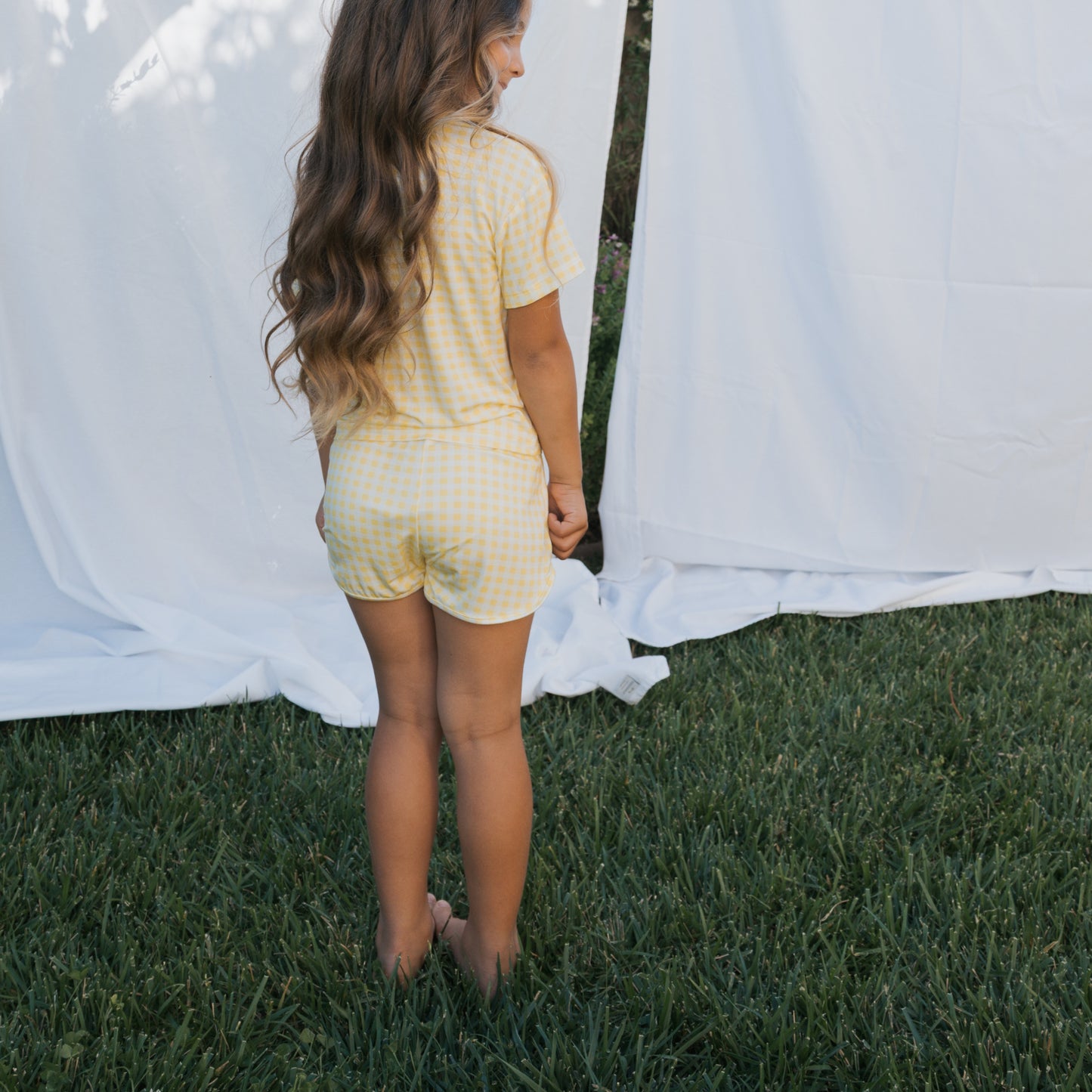 A young girl with long wavy hair stands barefoot on grass, facing away from the camera. She wears the forever french baby Girl's Bamboo Short Set in Citron, a yellow checkered breathable outfit. White sheets hang in the background.