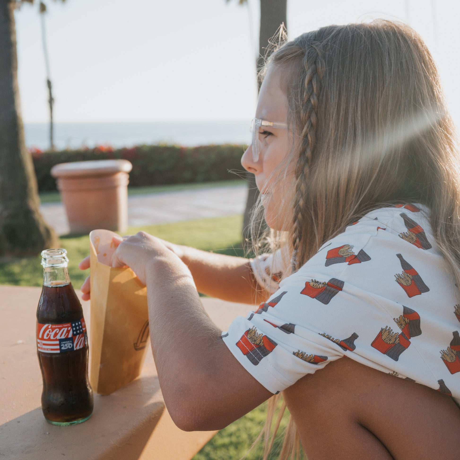 A young girl in the forever french baby Girl's Bamboo Short Set | Fries & Cola enjoys a sunny day at an outdoor table, reaching into a paper bag with a Coca-Cola bottle beside her. She looks comfy in breathable bamboo as palm trees sway in the background.