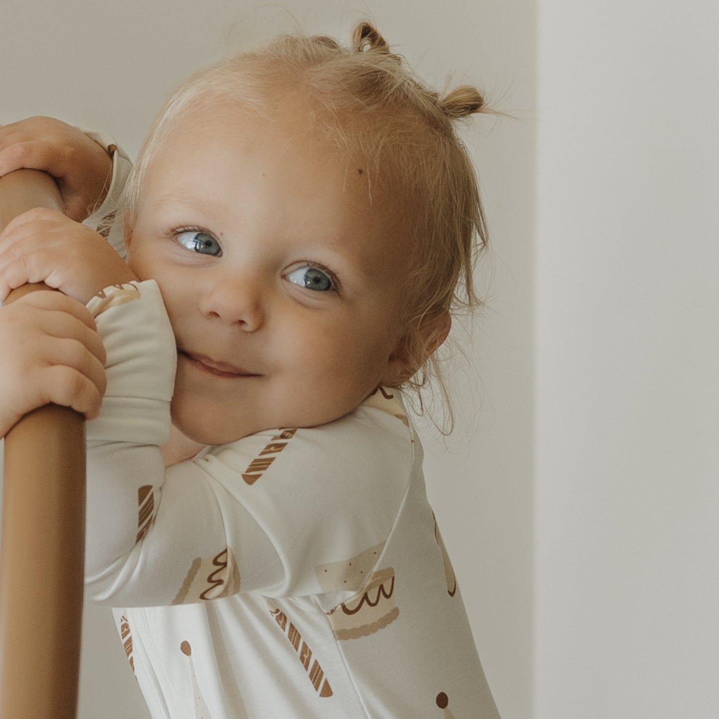 A young child with blonde hair in small pigtails smiles and hugs a wooden railing, wearing the forever french baby Bamboo Zip Pajamas | Birthday Girl—cream-colored hypoallergenic pajamas featuring brown dessert designs—against a neutral background.