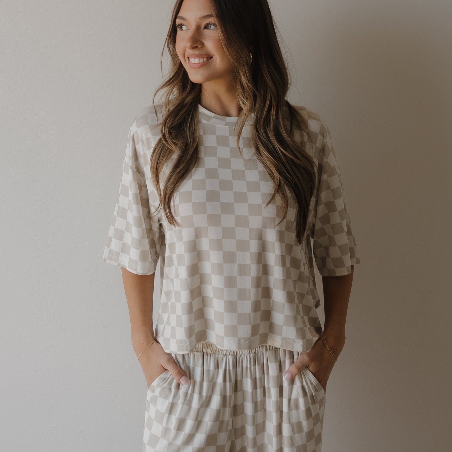 A woman stands against a light wall, smiling to the side, wearing the forever french baby Women's Boxy Tee Lounge Set | Checkerboard in beige and white, with her hands in her pockets.