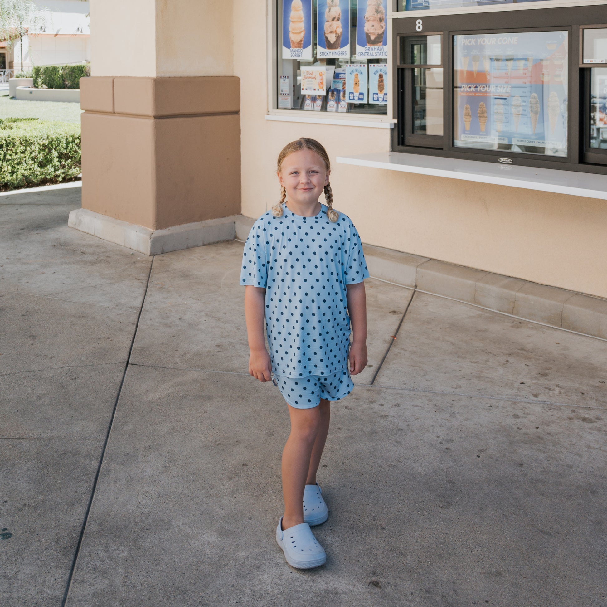 A young girl wearing the forever french baby Girl's Bamboo Short Set | Sully stands outside an ice cream shop, smiling at the camera. The shop window and menu are visible in the background.
