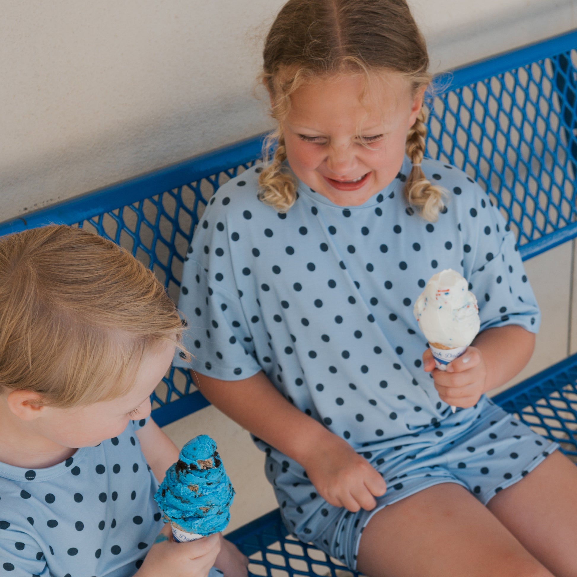 Two children in matching, breathable blue polka dot outfits sit on a blue metal bench, holding ice cream cones. The girl, dressed in the forever french baby Girl’s Bamboo Short Set | Sully, looks upset as her melting ice cream drips onto her hand, while the boy smiles at his bright blue scoop.