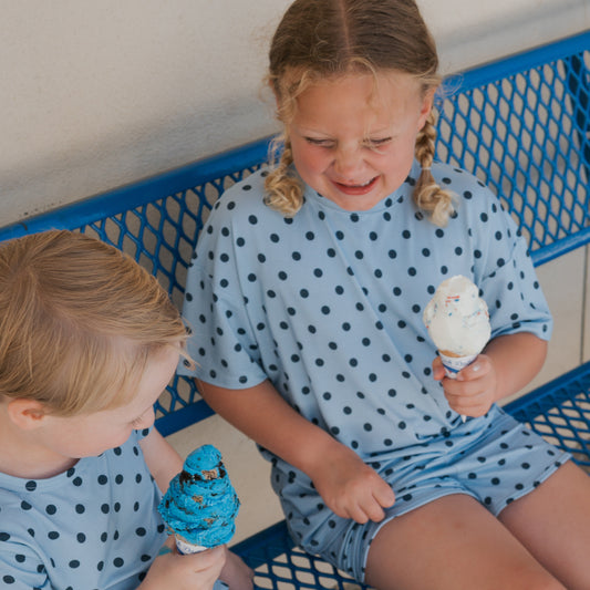 Two children in matching, breathable blue polka dot outfits sit on a blue metal bench, holding ice cream cones. The girl, dressed in the forever french baby Girl’s Bamboo Short Set | Sully, looks upset as her melting ice cream drips onto her hand, while the boy smiles at his bright blue scoop.