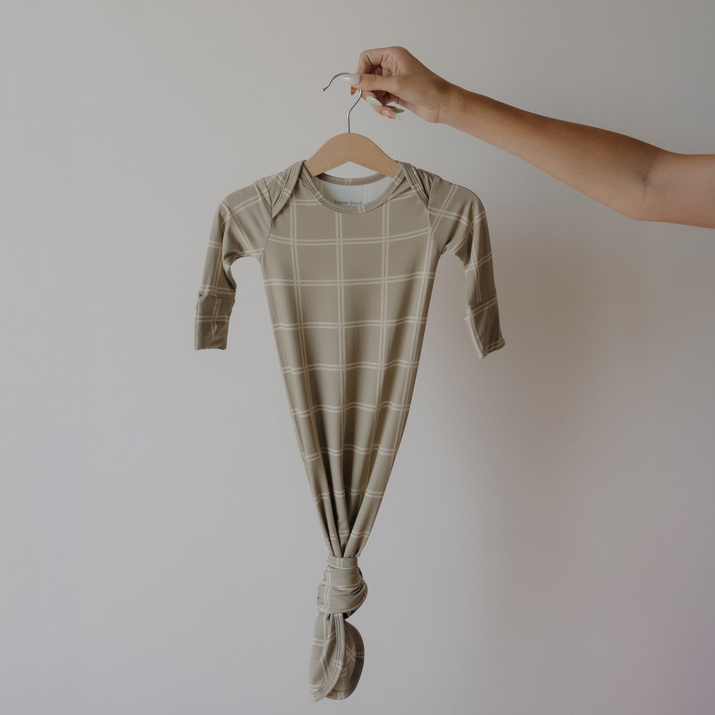 A hand holds the forever french baby Bamboo Knotted Gown | Linen Grid, a beige, long-sleeved bamboo baby gown with a white plaid pattern and knotted bottom—perfect as a newborn essential—against a plain white background.