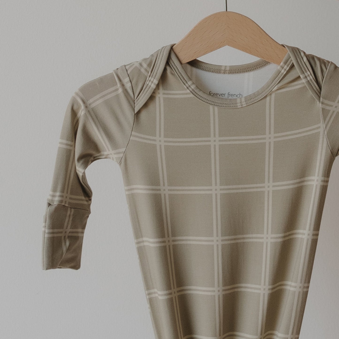 The Bamboo Knotted Gown | Linen Grid by forever french baby, featuring a light plaid pattern in beige, hangs on a wooden hanger—an ultra-cozy, hypoallergenic addition to your newborn essentials.