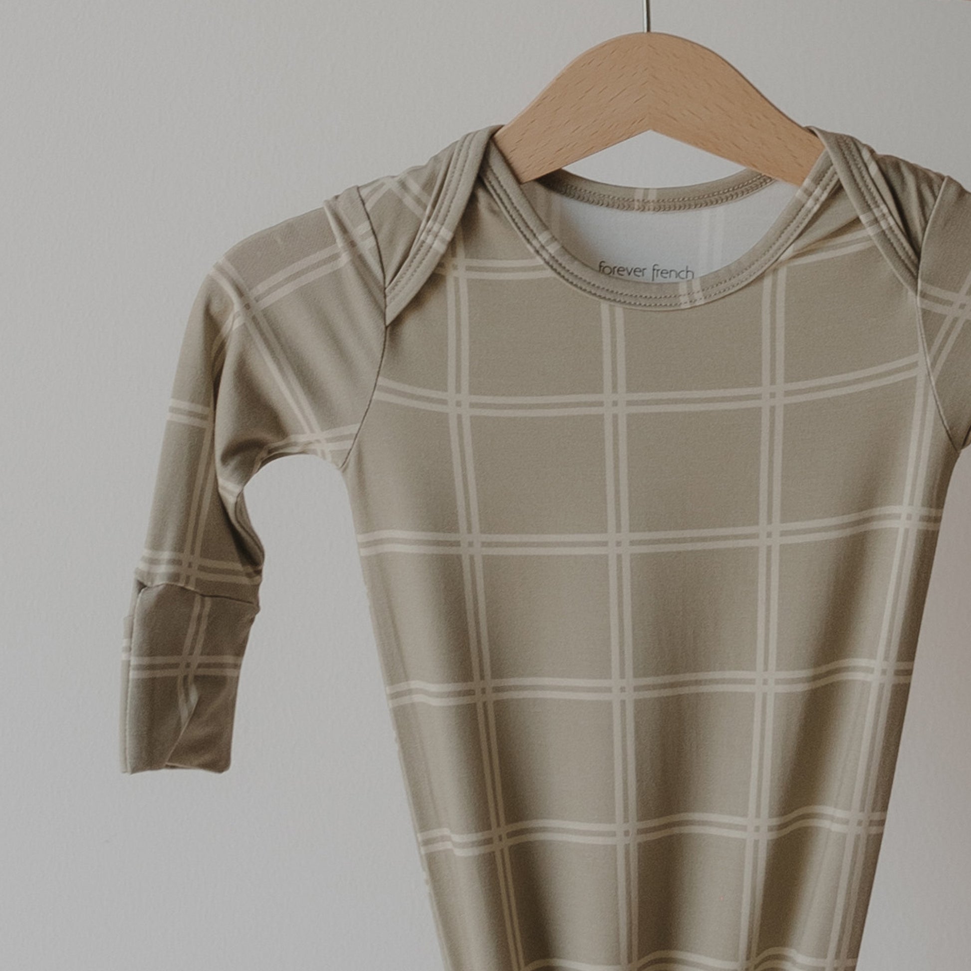 The Bamboo Knotted Gown | Linen Grid by forever french baby, featuring a light plaid pattern in beige, hangs on a wooden hanger—an ultra-cozy, hypoallergenic addition to your newborn essentials.