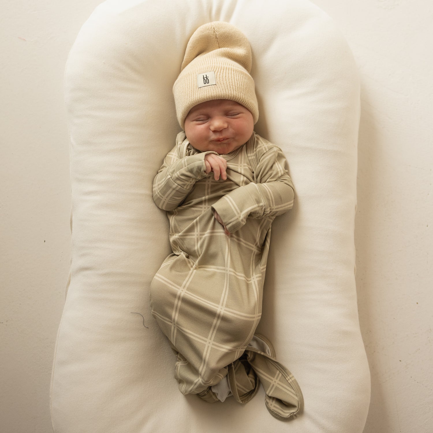 A newborn sleeps peacefully on a white cushion, wrapped in the forever french baby Bamboo Knotted Gown | Linen Grid, which keeps their hands snug and face calm. The hypoallergenic bamboo fabric ensures comfort and coziness.