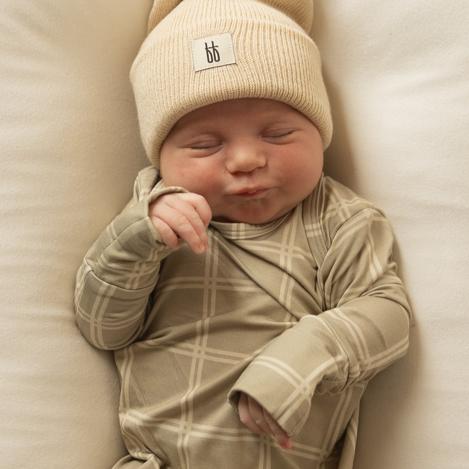 A newborn sleeps peacefully on a soft white surface, smiling sweetly in the forever french baby Bamboo Knotted Gown | Linen Grid with a matching beanie—adorably comfy in hypoallergenic bamboo clothing.