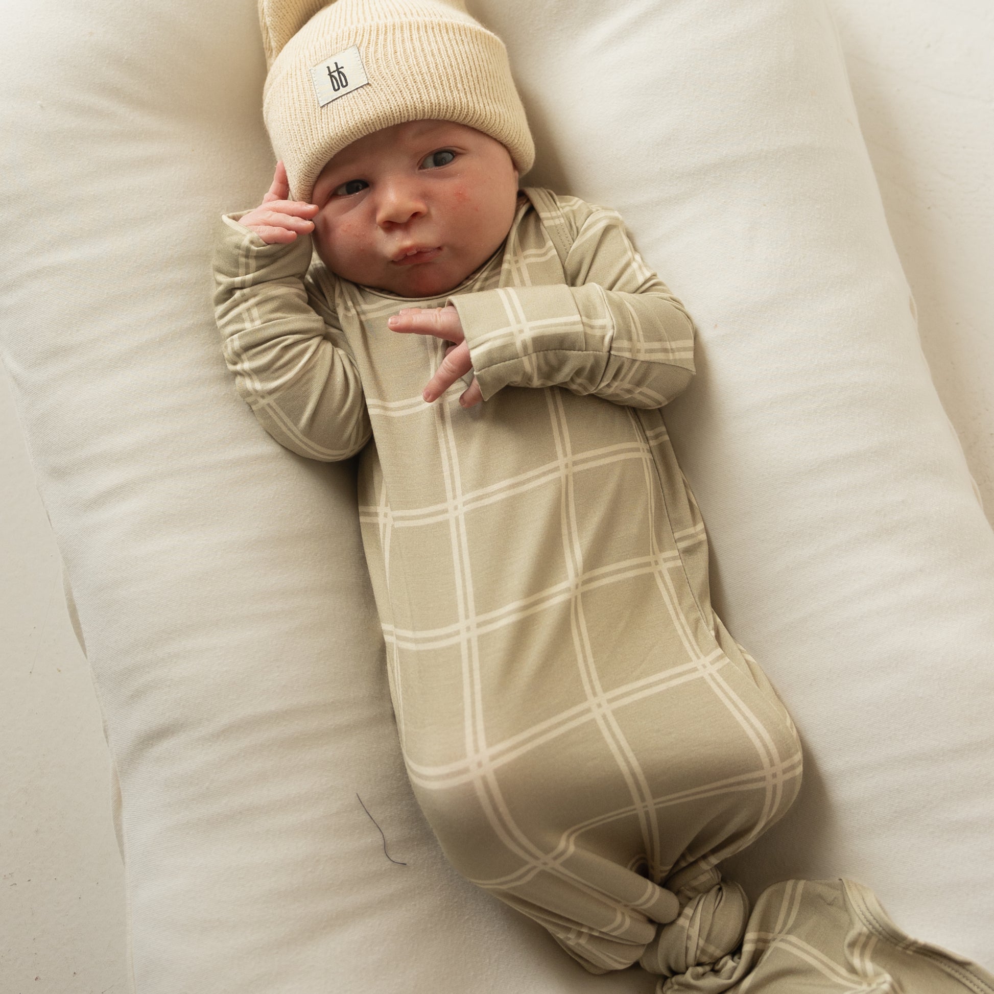 A newborn baby in the forever french baby Bamboo Knotted Gown | Linen Grid and a soft knit beanie lies on a white cushion, gazing upward with one hand near their face.