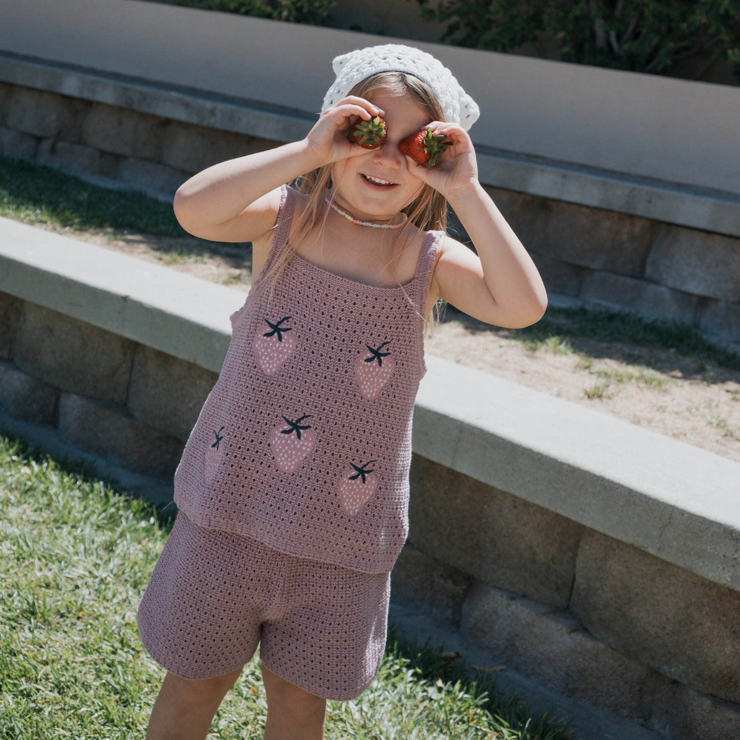 A young girl, smiling outdoors on the grass, playfully holds strawberries over her eyes while wearing the Matilda Girl's Knit Short Set by forever french baby and a white crocheted hat.