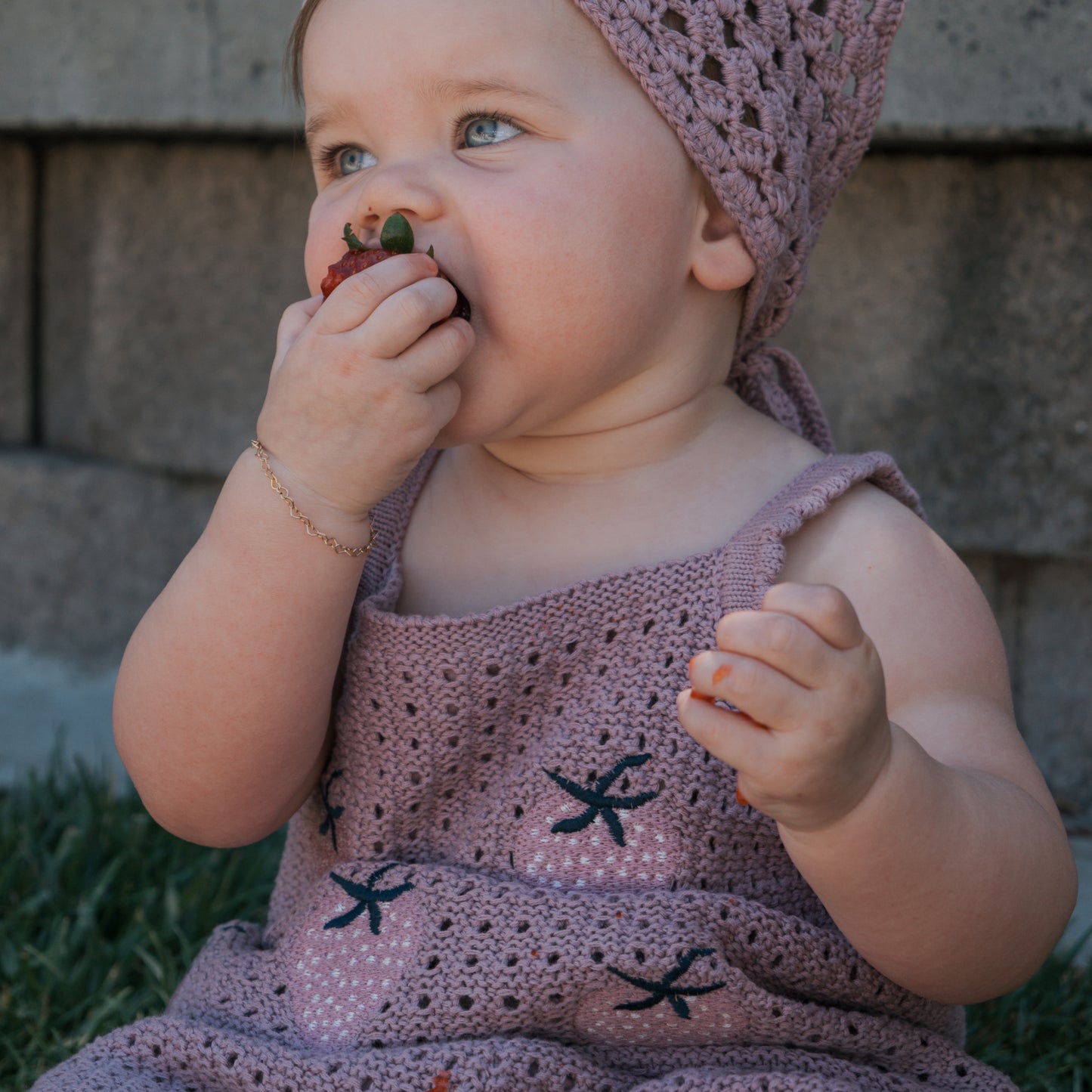 A baby wearing the forever french baby Girl's Knit Short Set | Matilda in purple sits outside, holding and eating a strawberry, with a brick wall in the background.