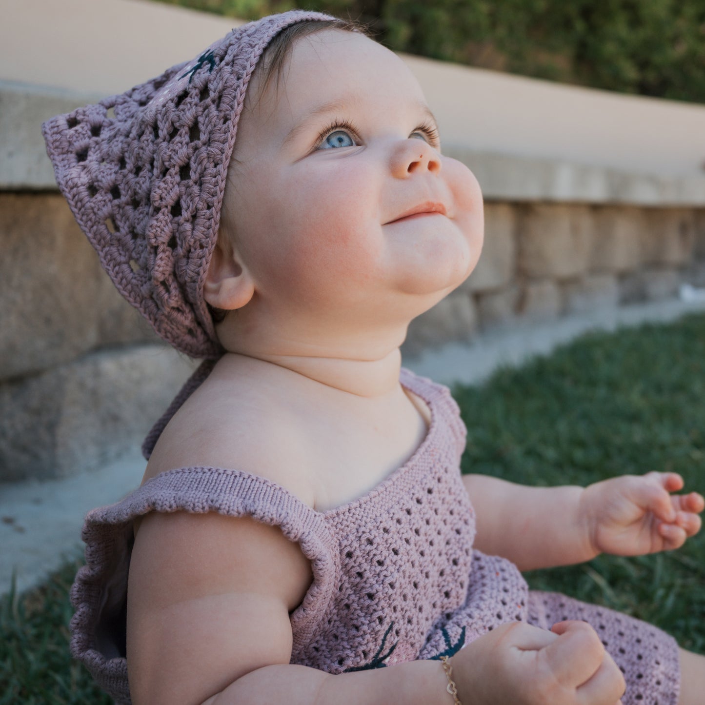 A baby wearing the Girl's Knit Short Set | Matilda by forever french baby sits on grass, looking up and smiling, with a stone wall and greenery in the background.
