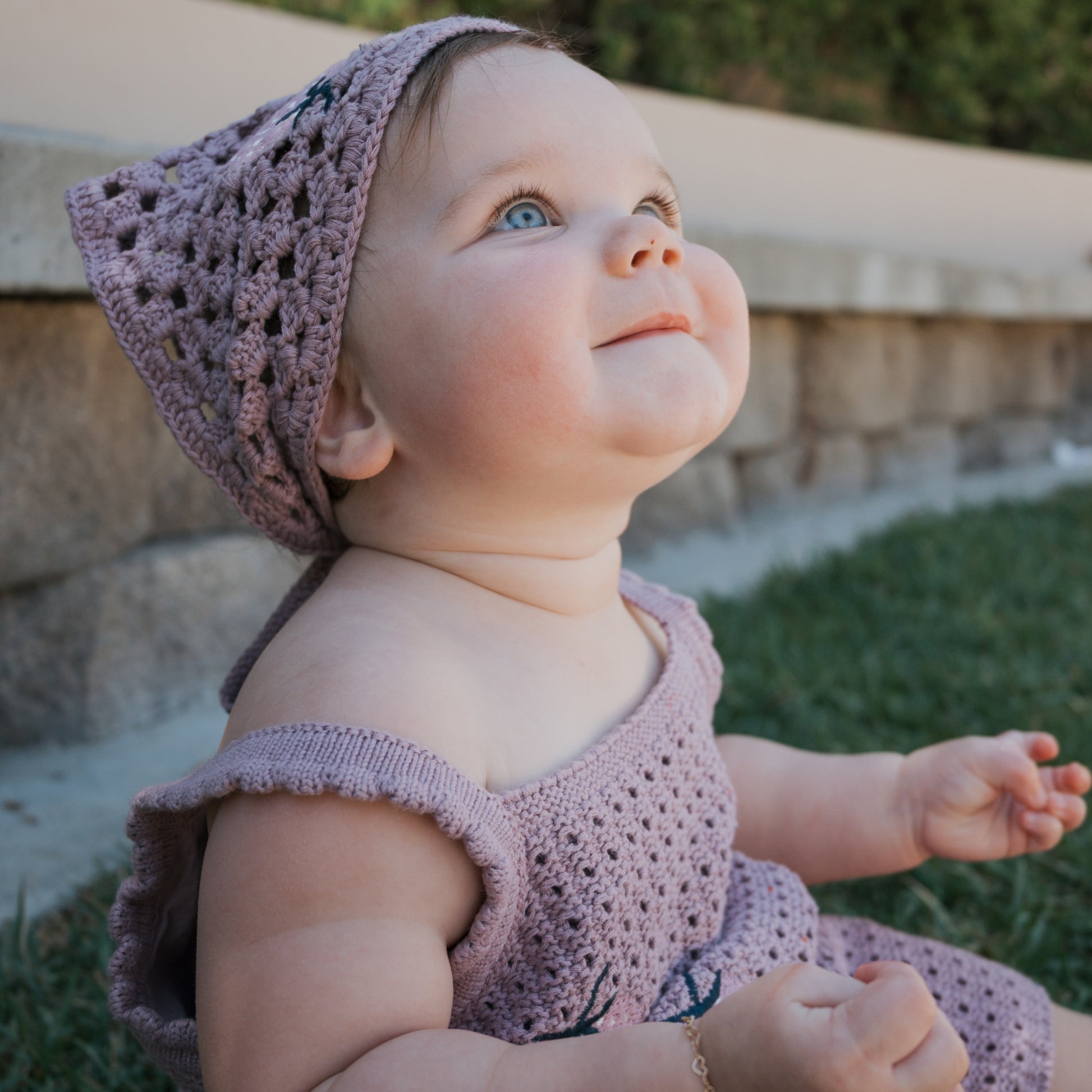 A baby wearing the Girl's Knit Short Set | Matilda by forever french baby sits on grass, looking up and smiling, with a stone wall and greenery in the background.