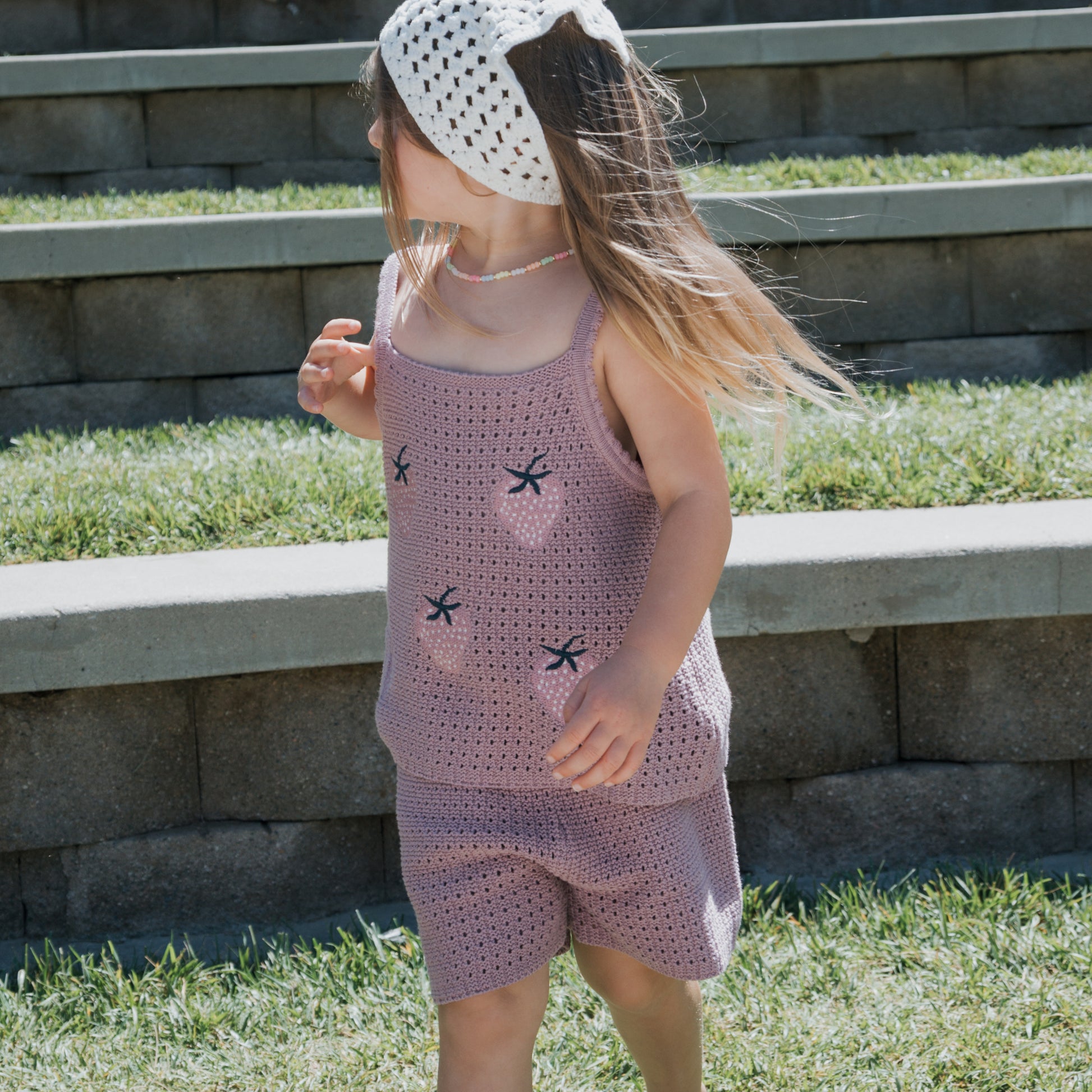 A young child wearing the forever french baby Girl's Knit Short Set | Matilda, a soft lavender 100% cotton crochet tank and shorts ensemble, paired with a white crochet headband and a beaded necklace, stands outdoors in front of stone steps on a sunny day.