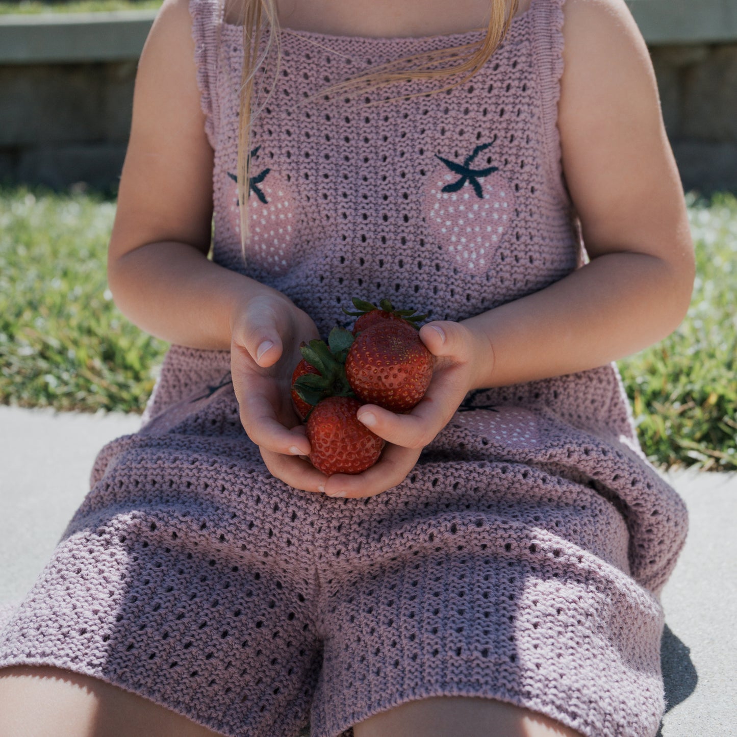 A child dressed in the Matilda Girl's Knit Short Set from forever french baby sits on the ground, holding fresh strawberries in both hands. The soft knit fabric enhances the playful scene, with grass and a stone wall visible in the background.