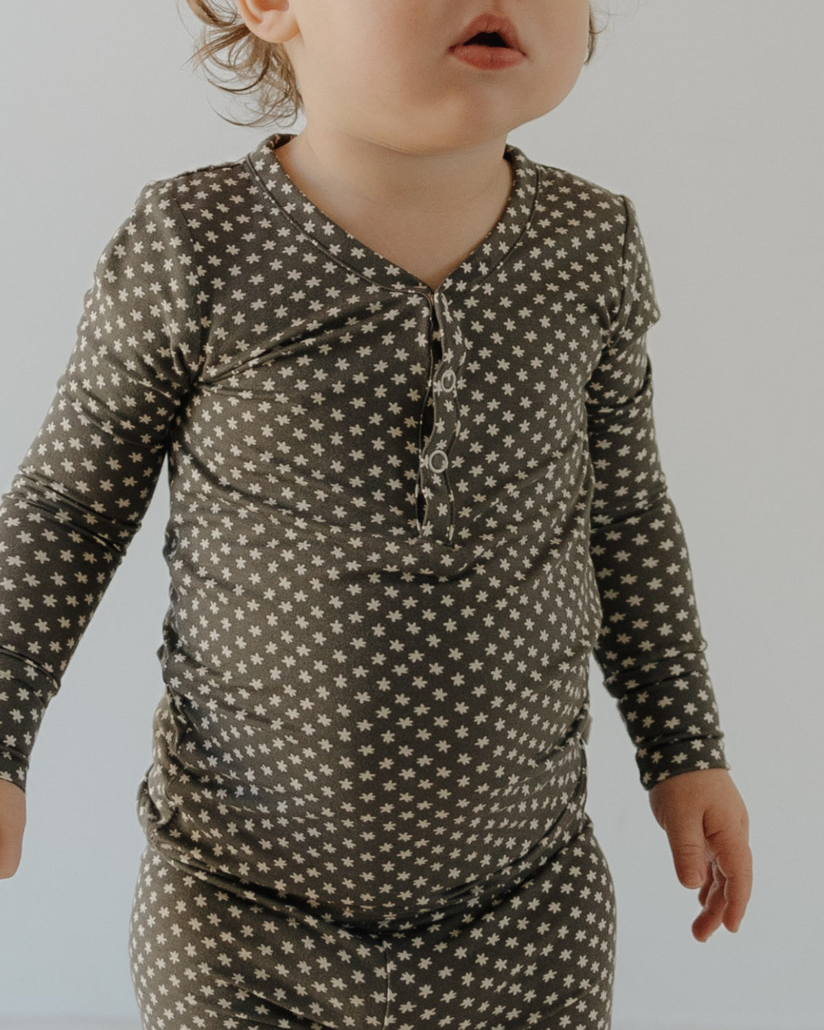 A young child in forever french baby Bamboo Two Piece Pajamas | Green Flowers—a long-sleeve, button-up kids sleepwear set—stands by a plain light wall, their face partially out of frame.