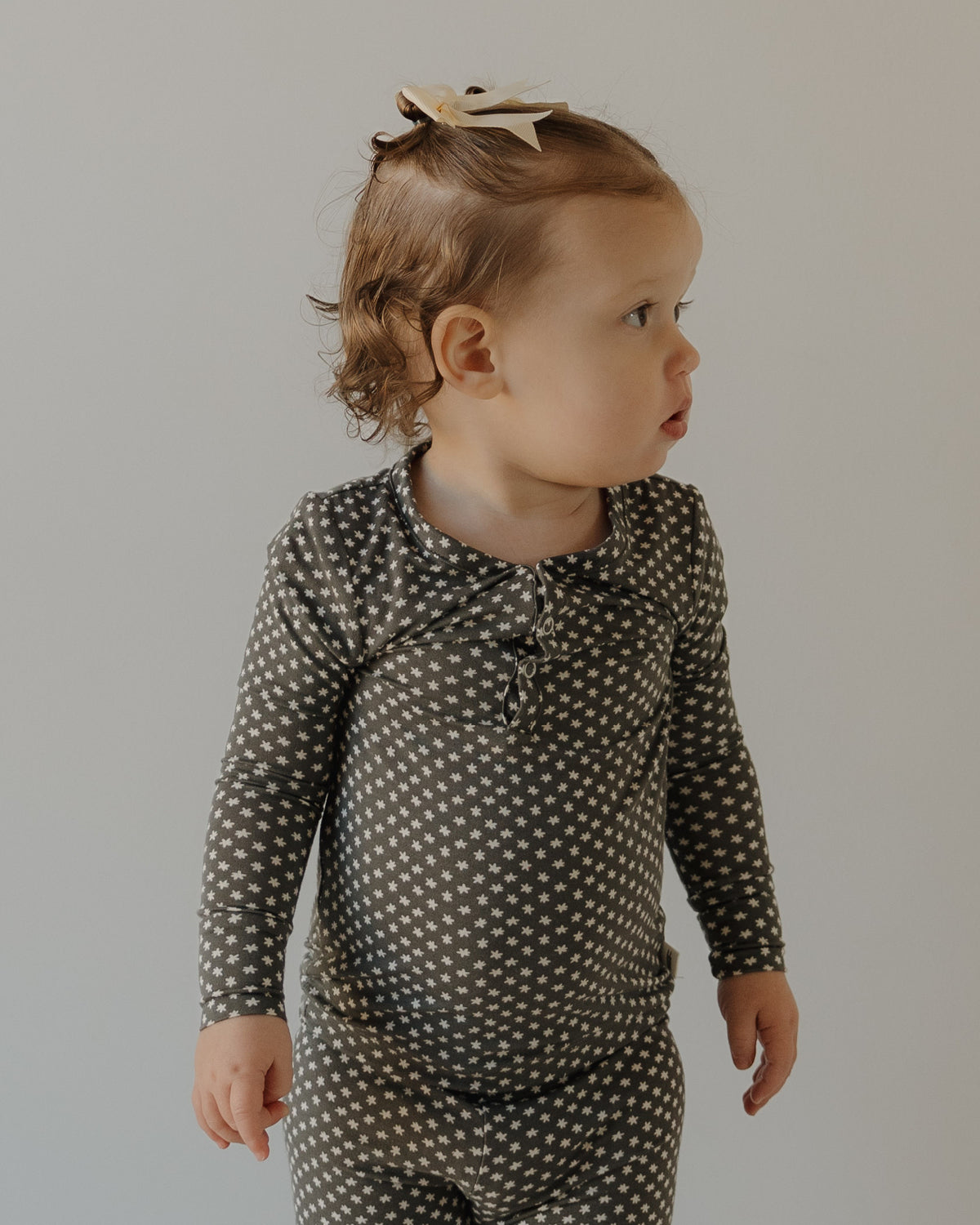 A young child with light brown hair in a bow stands against a plain background, wearing forever french baby's Bamboo Two Piece Pajamas in Green Flowers—breathable kids sleepwear perfect for comfort and style.