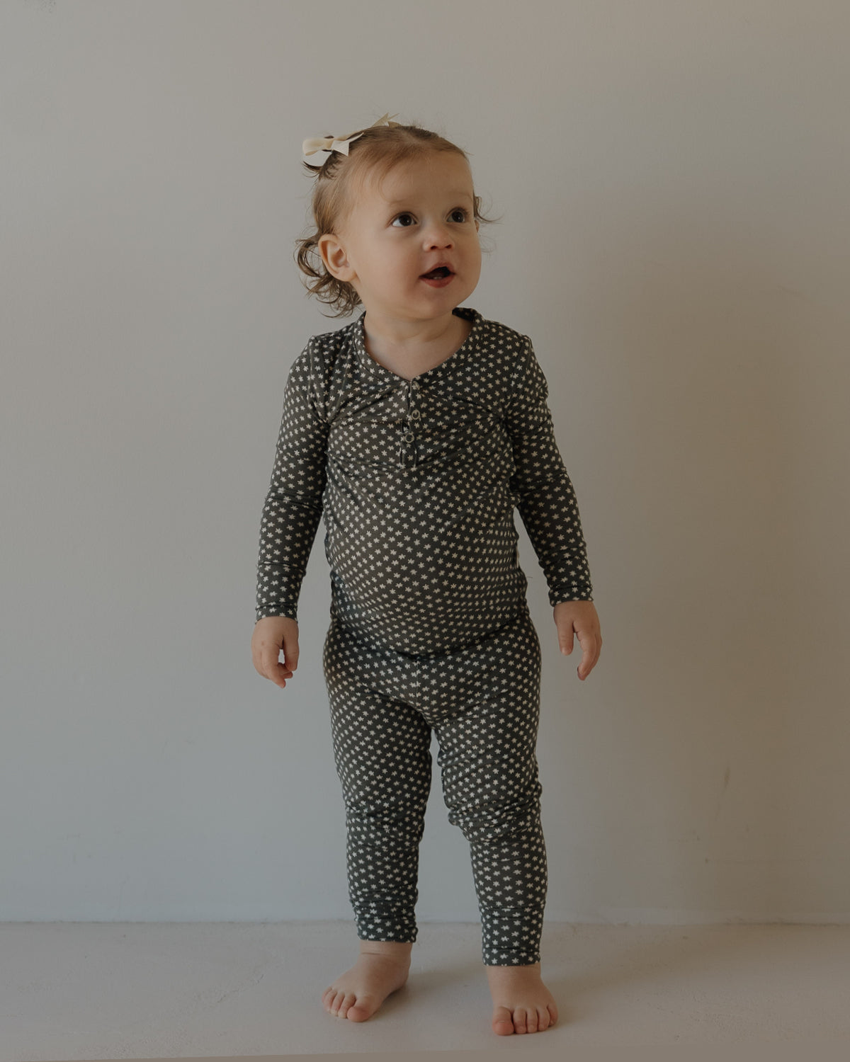 A toddler with light brown hair and a white bow stands barefoot against a light wall, wearing forever french baby Bamboo Two Piece Pajamas in Green Flowers. She looks up curiously, highlighting the comfy, breathable kids sleepwear.